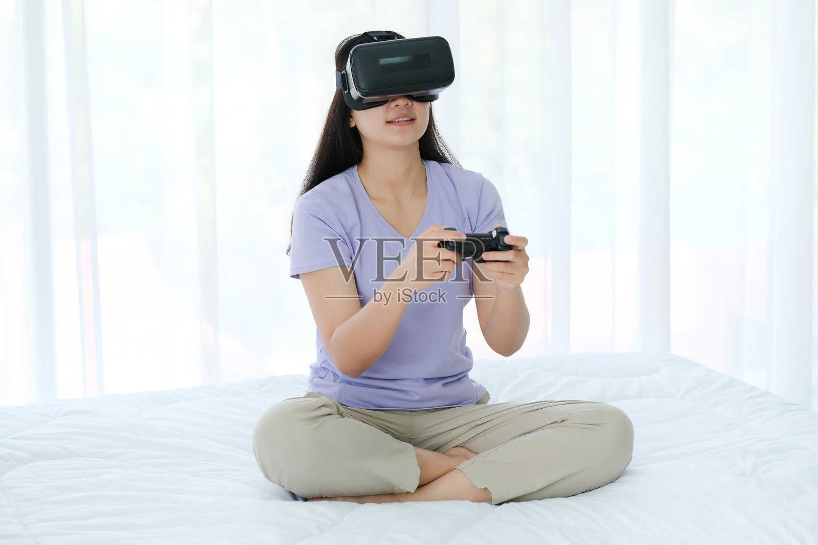 亚洲女性戴着虚拟现实眼镜，坐在卧室的床上。手持操纵杆，使用VR眼镜玩游戏照片摄影图片