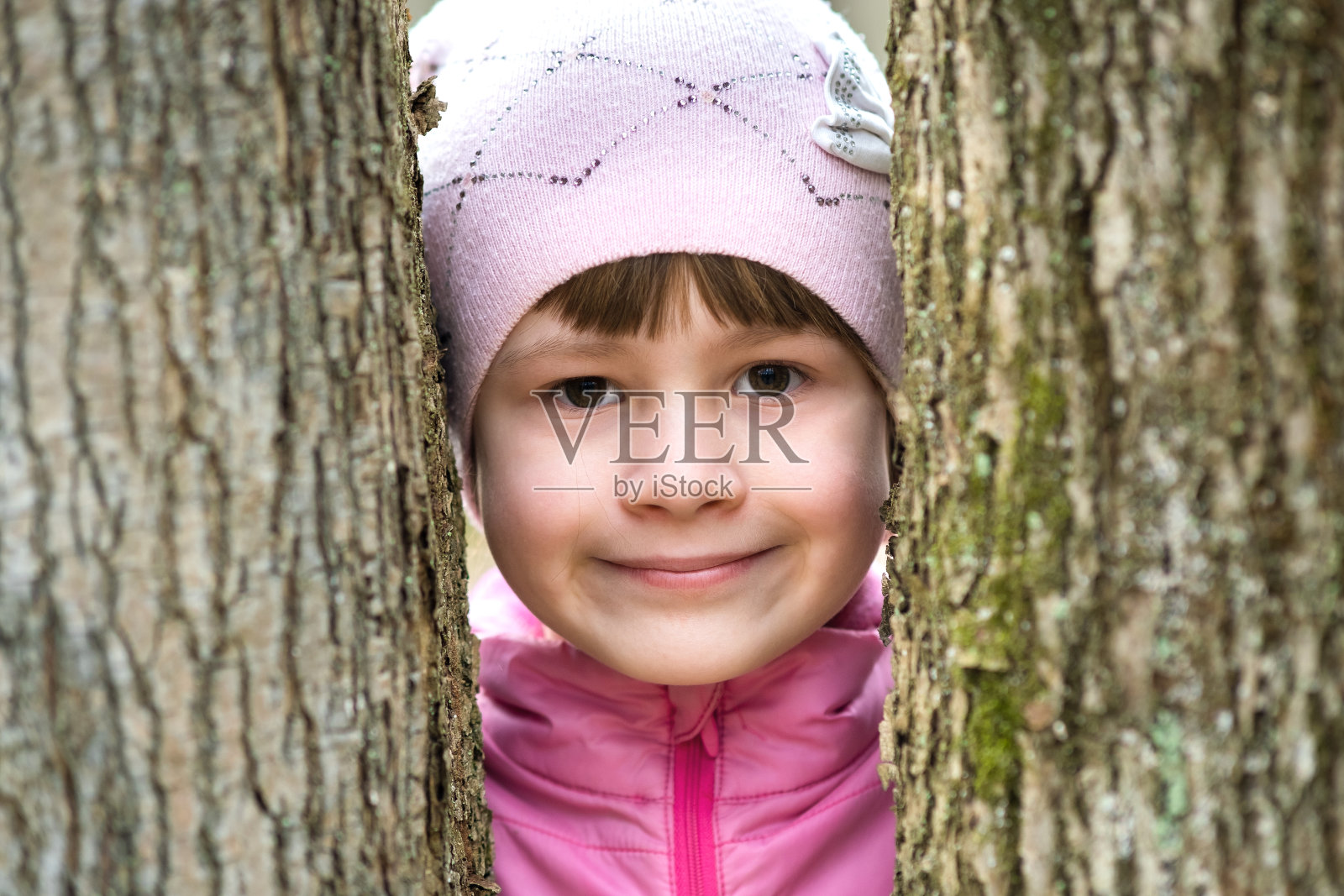 年轻漂亮的小女孩的肖像穿着粉红色的夹克和帽子站在树木之间的公园享受温暖的阳光天在早春户外。照片摄影图片