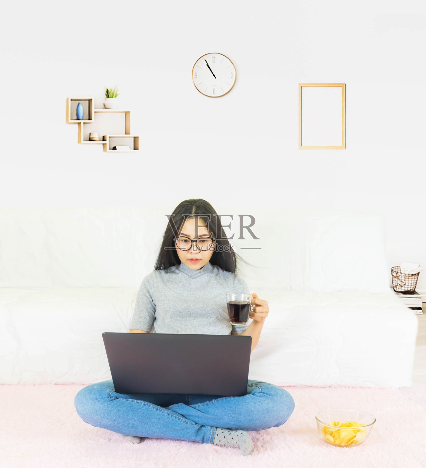 年轻美丽的亚洲女子穿着灰色t恤和牛仔裤，坐在客厅的粉色地毯上，在线工作，用咖啡杯在家放松，在家工作笔记本电脑，在家工作照片摄影图片