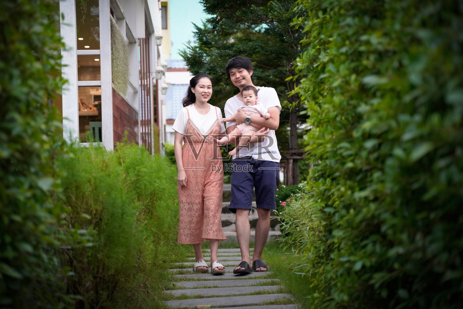 快乐年轻的亚洲家庭漫步在小花园的台阶上。父亲抱着7个月大的婴儿。每个人都很高兴在模糊的咖啡馆餐厅背景。照片摄影图片