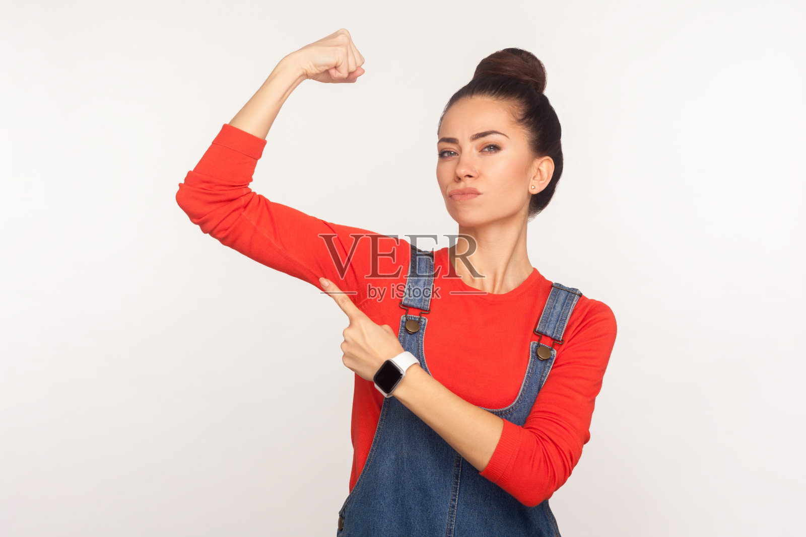 看，我是独立和坚强!一个自信的充满活力的时尚女孩在牛仔工装裤举起手指着二头肌照片摄影图片