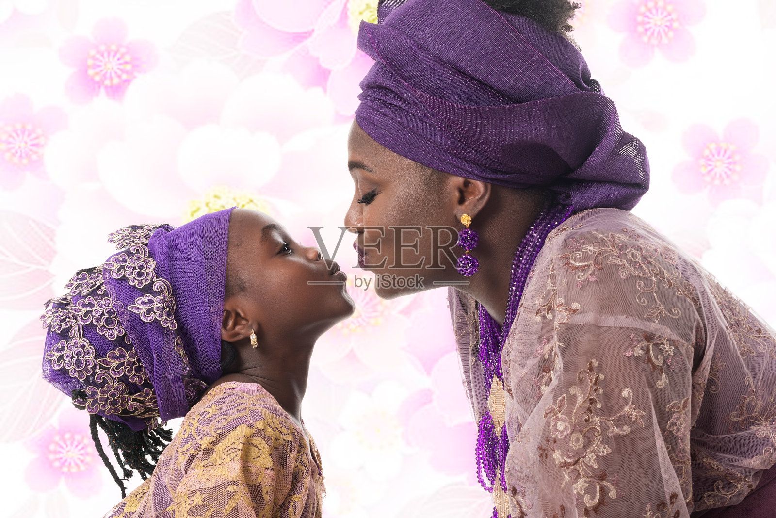 肖像在侧面的母亲和孩子女孩亲吻。以花朵为背景的非洲传统紫色服装照片摄影图片