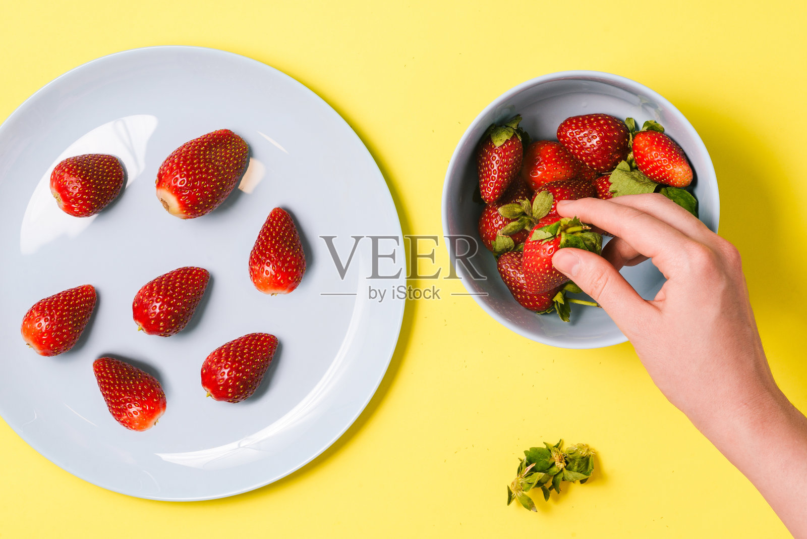 女孩的手拿着黄色背景上的草莓。配草莓的蓝色盘子。健康的夏季早餐，午餐或晚餐。俯视图，平放。照片摄影图片