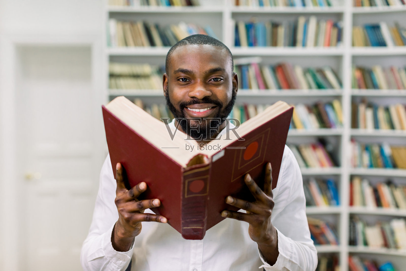 好看的满意的年轻非洲胡子男孩在白色休闲衬衫，摆姿势在书架的背景在现代图书馆，拿着大打开红书。书日快乐，教育理念照片摄影图片