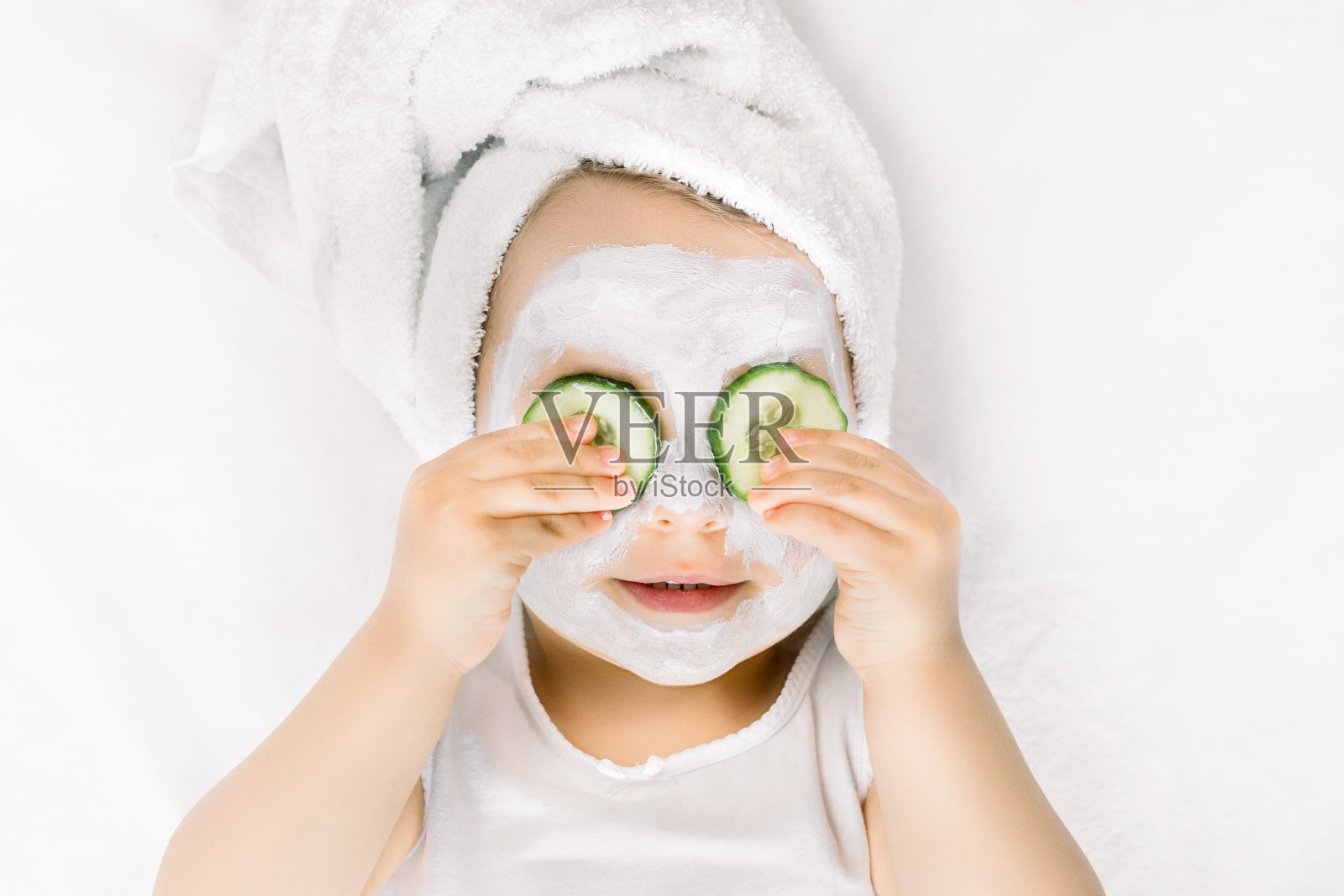 在家做面部水疗。特写滑稽可爱的小女孩在白色浴巾拿新鲜黄瓜片在她的眼睛躺在白色的背景照片摄影图片