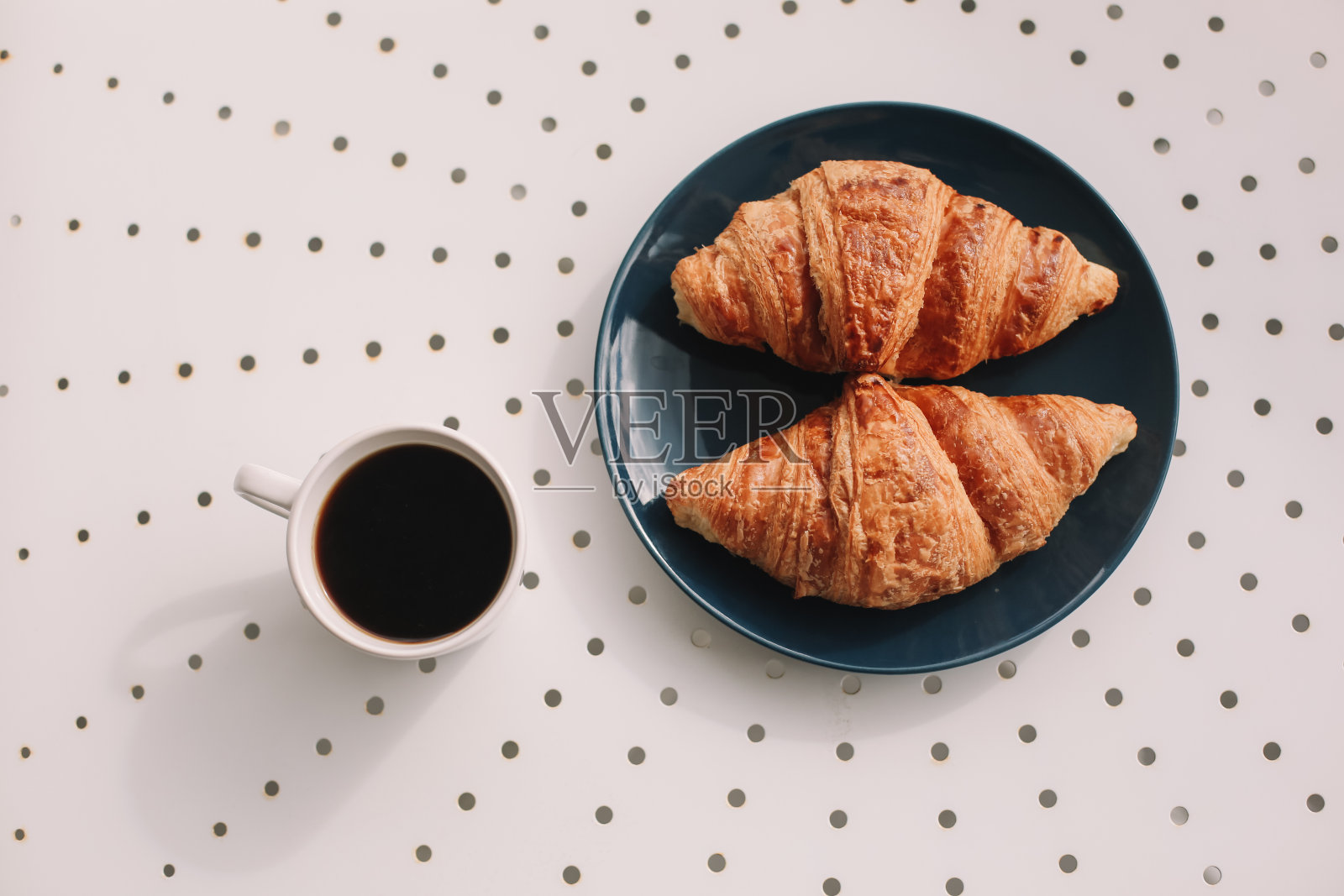 咖啡杯和新鲜出炉的法式羊角面包在蓝色的盘子和白色的背景。早餐。照片摄影图片
