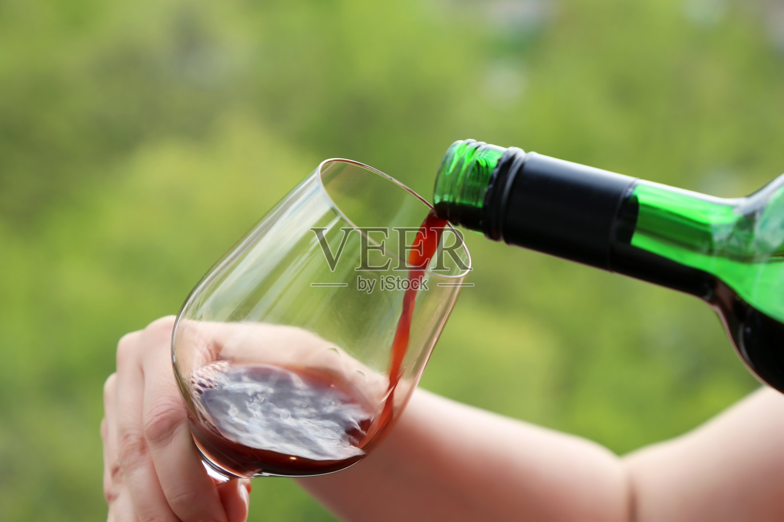 女子将酒瓶中的红酒倒进绿色自然背景的玻璃杯中照片摄影图片