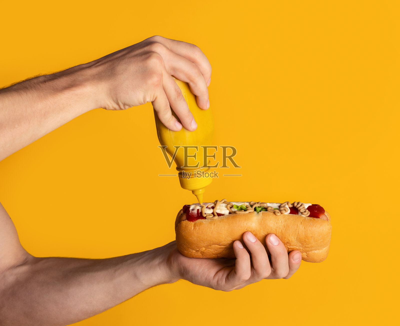 年轻人将芥末酱涂在新鲜多汁的热狗上，背景为橙色。特写镜头照片摄影图片