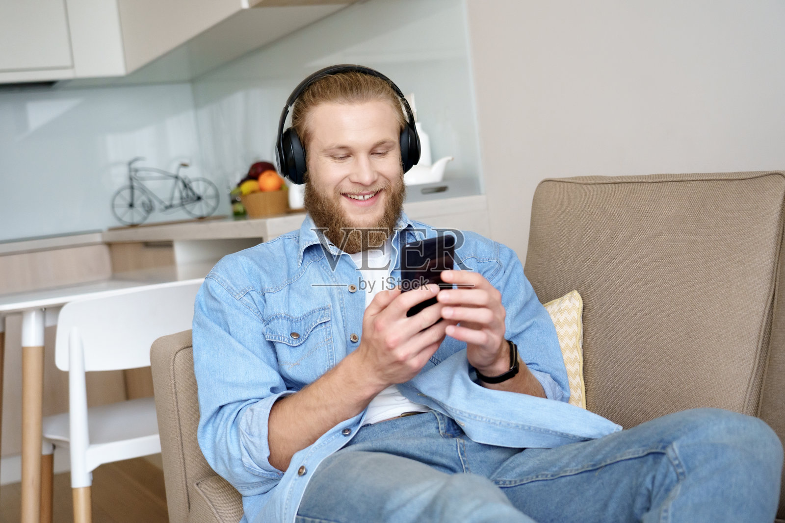 快乐的年轻人戴着无线耳机听音乐，有声书或学习语言的智能手机上的应用程序坐在沙发上。放松的人享受移动播放器播客休息室在沙发上在家。照片摄影图片