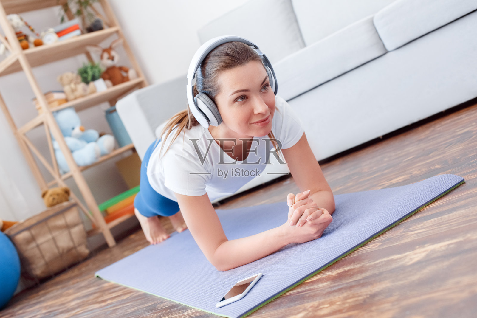 健康的生活方式。一名年轻女子戴着耳机在家里做瑜伽，做前臂平板支撑的姿势，听着智能手机上健身应用程序的说明，专注地微笑照片摄影图片