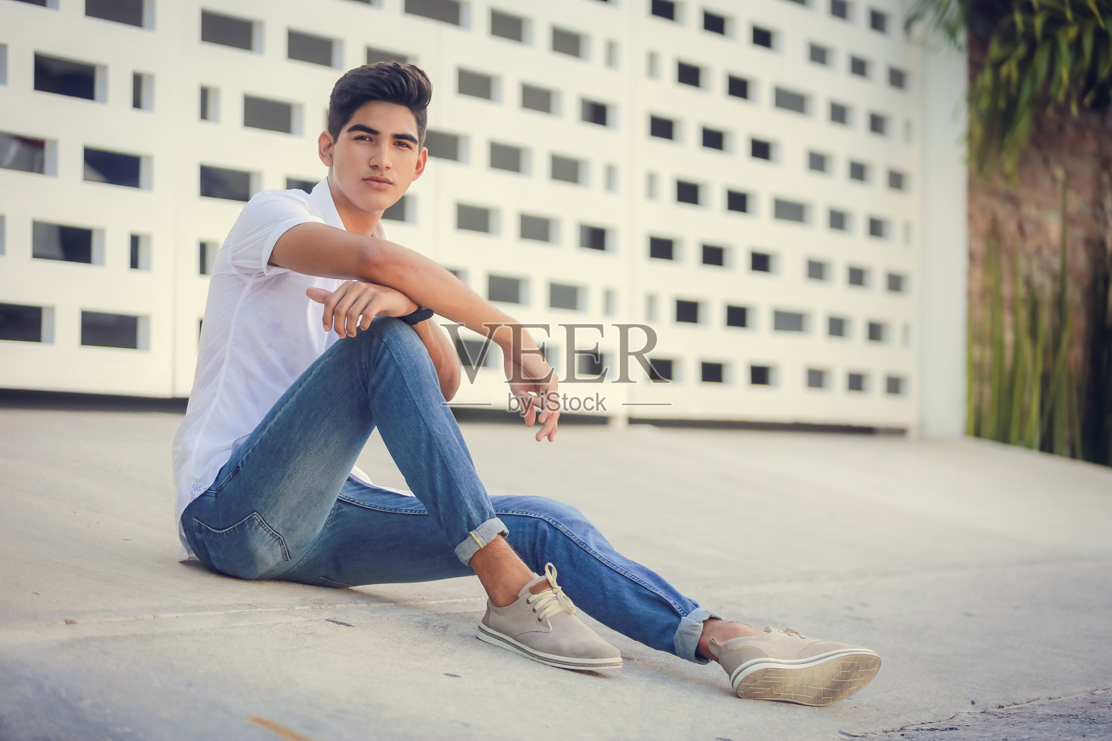 穿着牛仔裤和白衬衫的现代漂亮的年轻男子模特坐在城市夏季室外露台的地板上。有型有吸引力的西班牙潮男室外模特。照片摄影图片