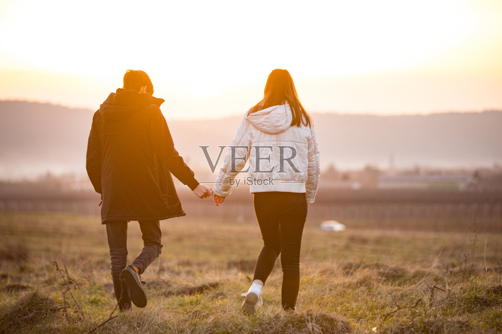 后视图的十几岁的夫妇牵着手走在草地在日落照片摄影图片
