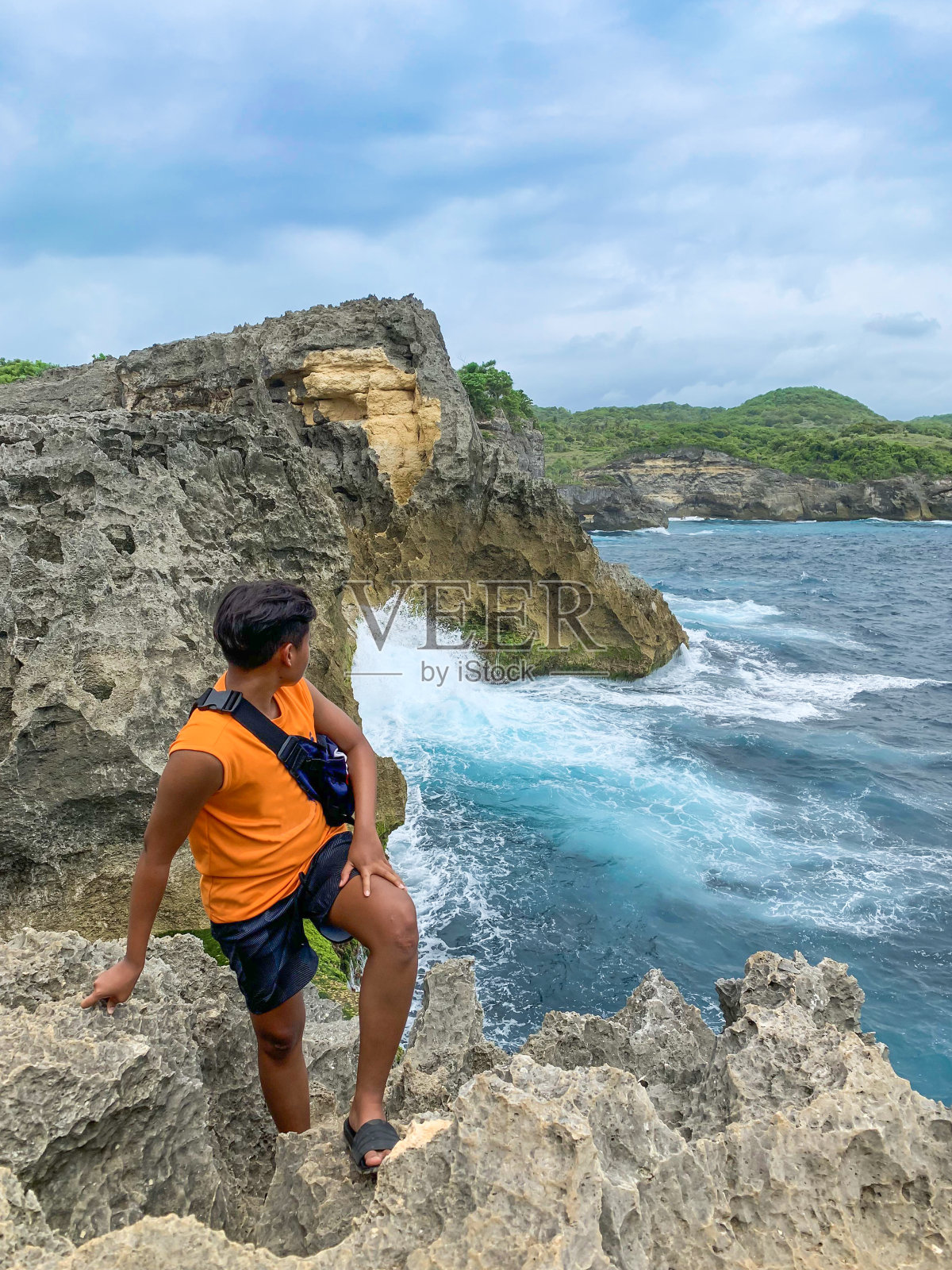 年轻的游客站在悬崖的边缘，回头看海岸边的巨石。海浪在海浪背景上撞击岩石。夏天的风景。重镇penida,巴厘岛照片摄影图片