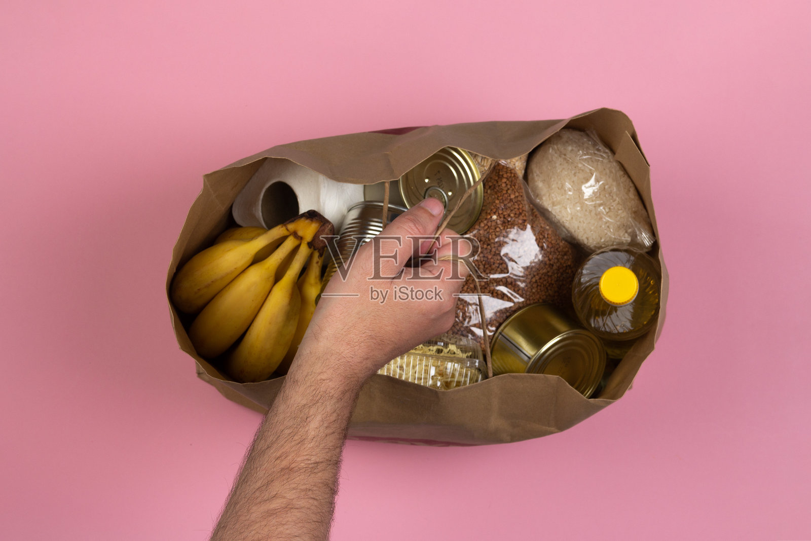 纸袋与危机食品供应隔离隔离期间的粉红色背景，意大利面，荞麦，罐头食品，大米，香蕉在一个男性的手特写。送餐、捐赠理念。照片摄影图片