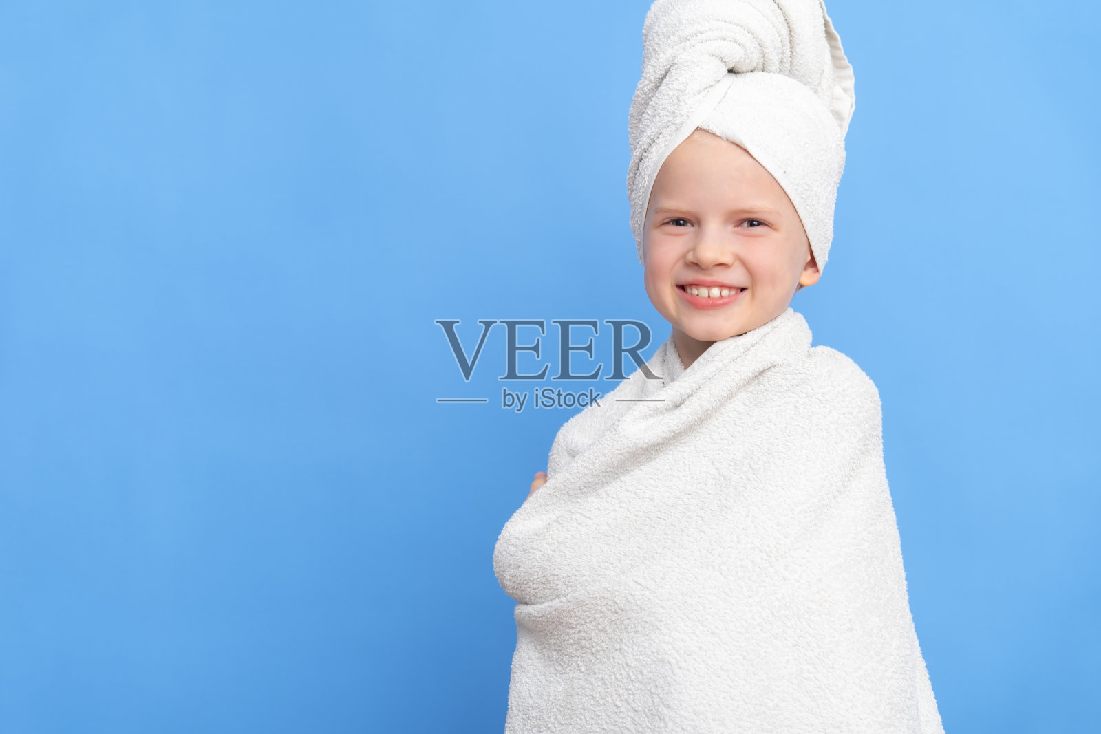 可爱的小白人女孩的肖像包裹在白色的毛巾与浴巾在头上的蓝色工作室背景。水疗程序，沐浴纺织品照片摄影图片