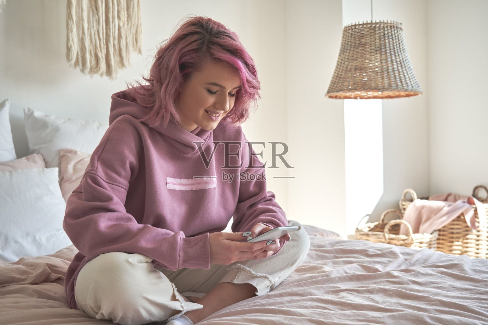粉色头发的时尚少女微笑着拿着智能手机在社交媒体上聊天，看视频内容，发短信，玩游戏，使用手机应用程序，上网坐在家里的床上。照片摄影图片