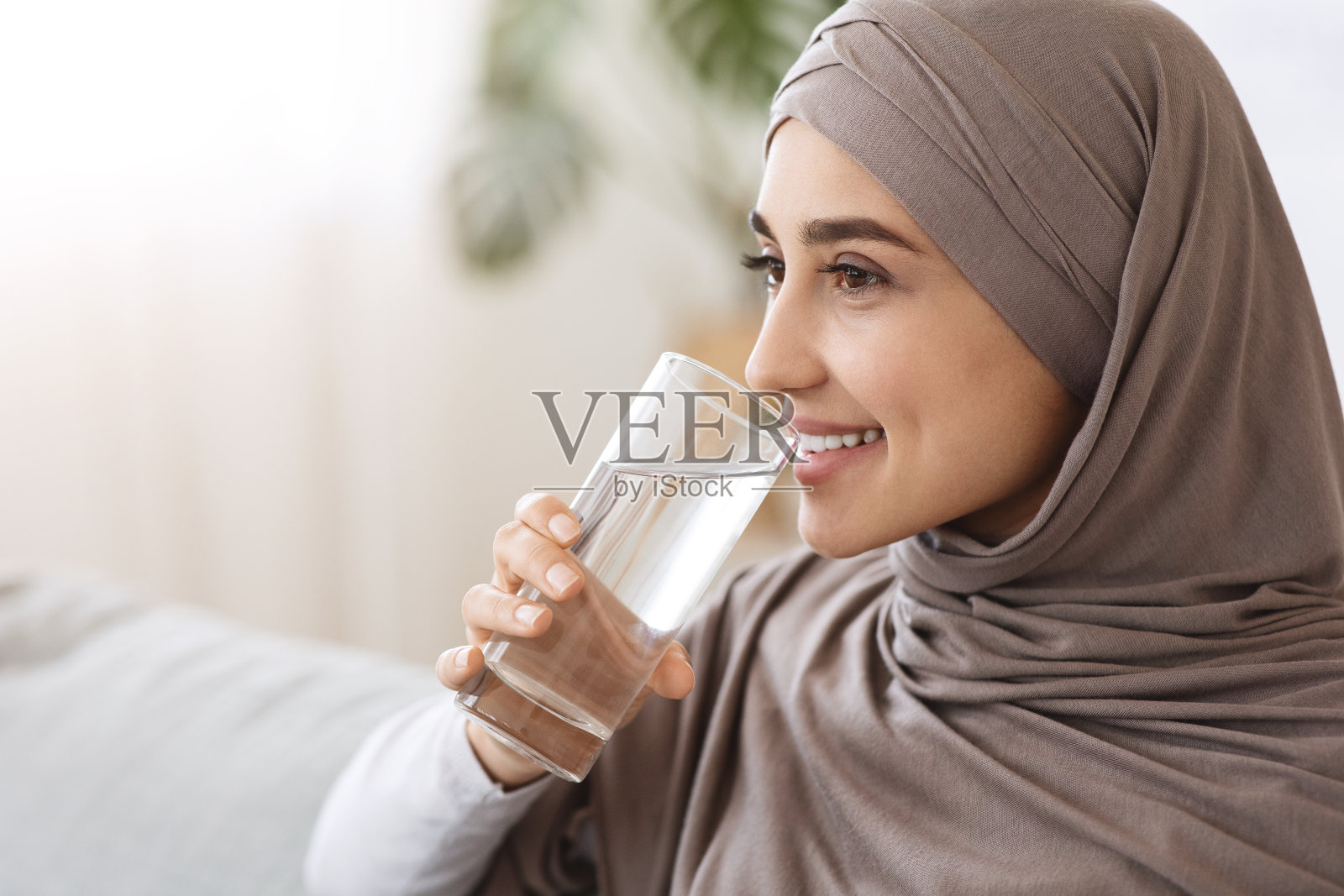 健康的液体。微笑的穆斯林妇女在家喝矿泉水，照片摄影图片