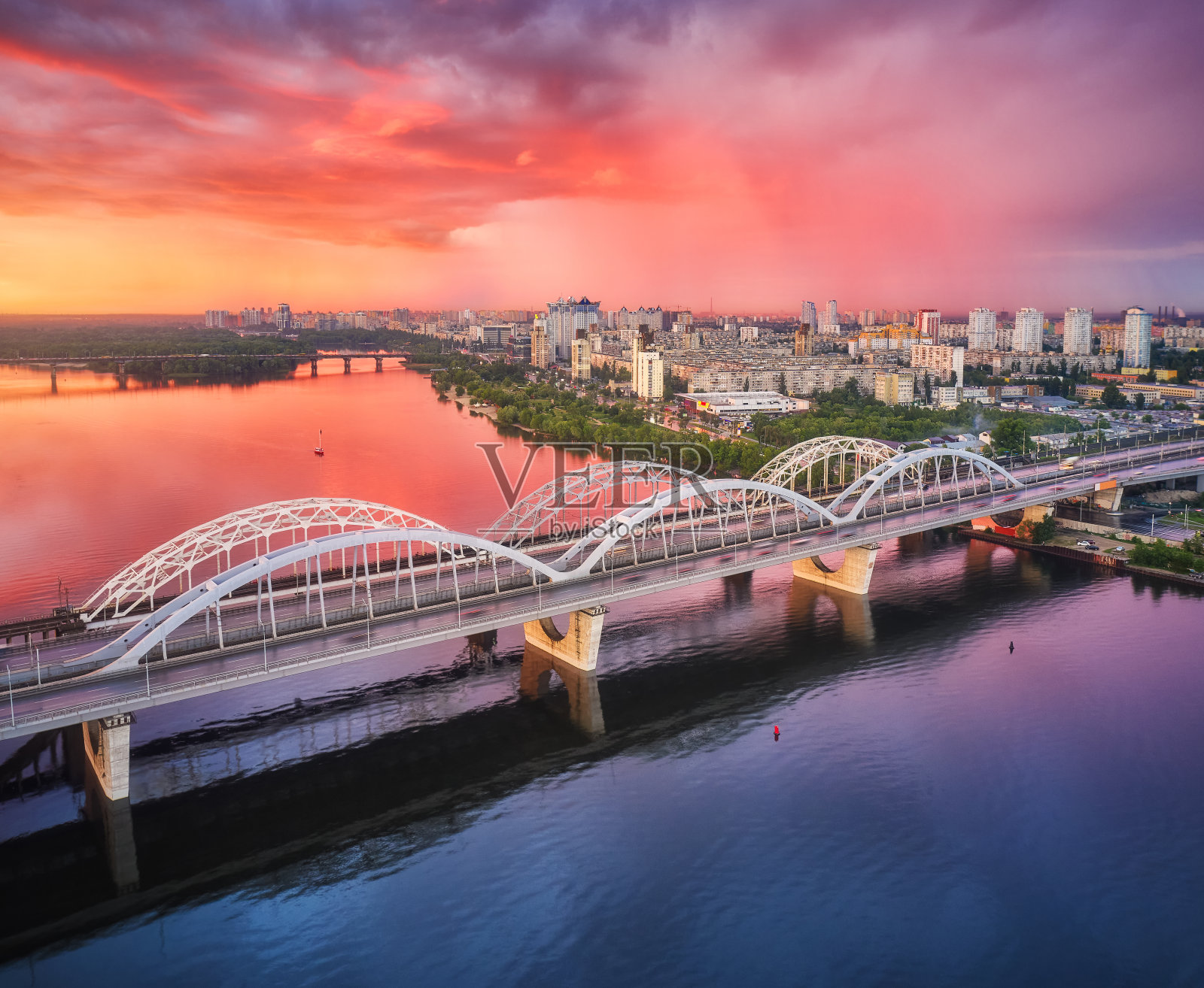 鸟瞰图美丽的桥在日落在基辅，乌克兰。风景有桥，河流，城市，彩色的天空和红色的云在夏天。城市景观与道路，建筑，倒影在水中。俯视图照片摄影图片