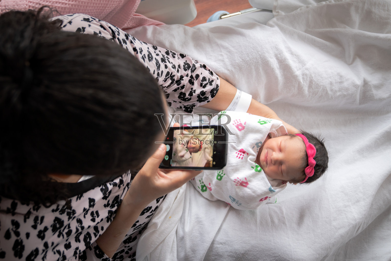 一位身穿粉色动物印花浴袍的母亲坐在医院的病床上，抱着襁褓中的新生女婴，用手机拍照，以便与家人和朋友分享。照片摄影图片