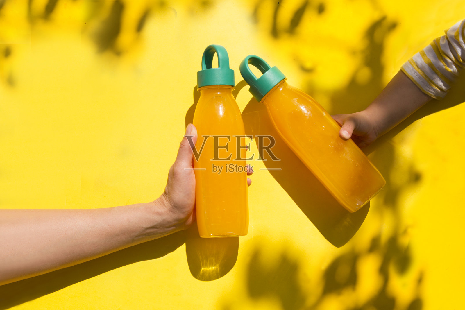 成人和儿童的手拿瓶黄色的夏季饮料:冰沙或果汁在黄色的背景与树影。夏天的概念。照片摄影图片