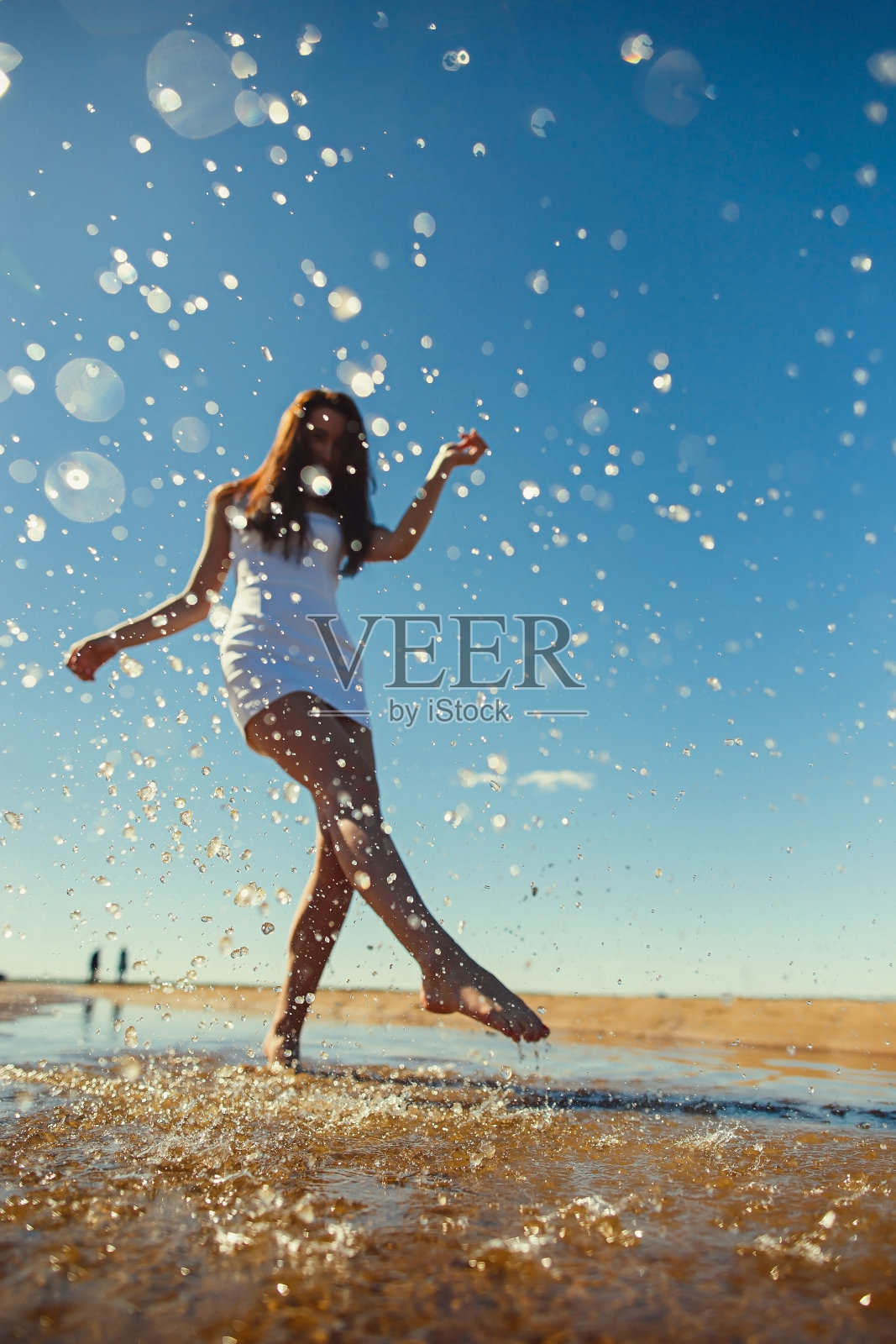 阳光下，一个穿着白色连衣裙的苗条女孩在水面上溅起水花。夏天在沙滩上休息的概念，好天气，快乐照片摄影图片