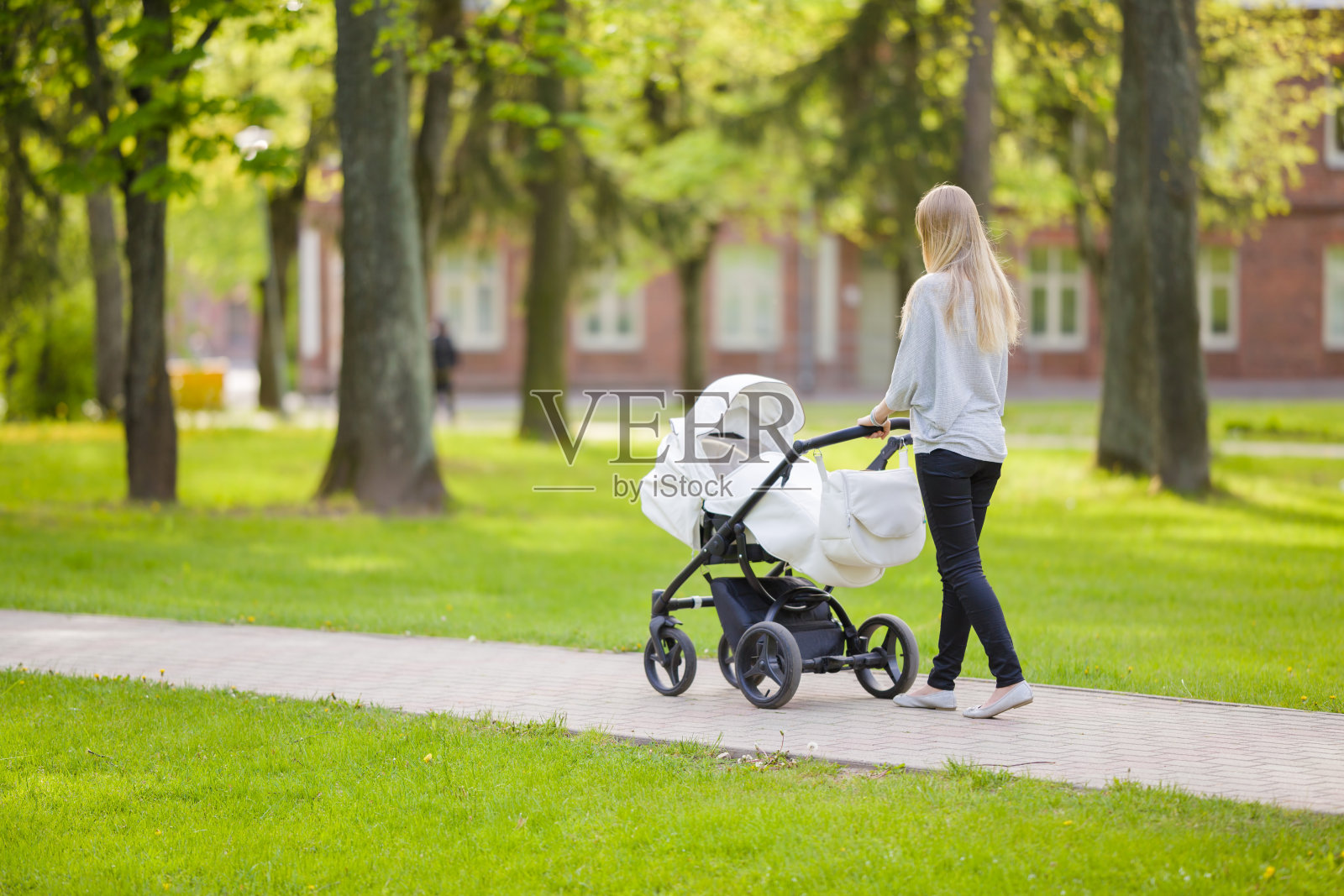 一位年轻的母亲推着一辆白色婴儿车，在温暖、阳光明媚的春天里，慢慢地走在镇绿公园里。花时间陪婴儿，呼吸新鲜空气。享受漫步。照片摄影图片