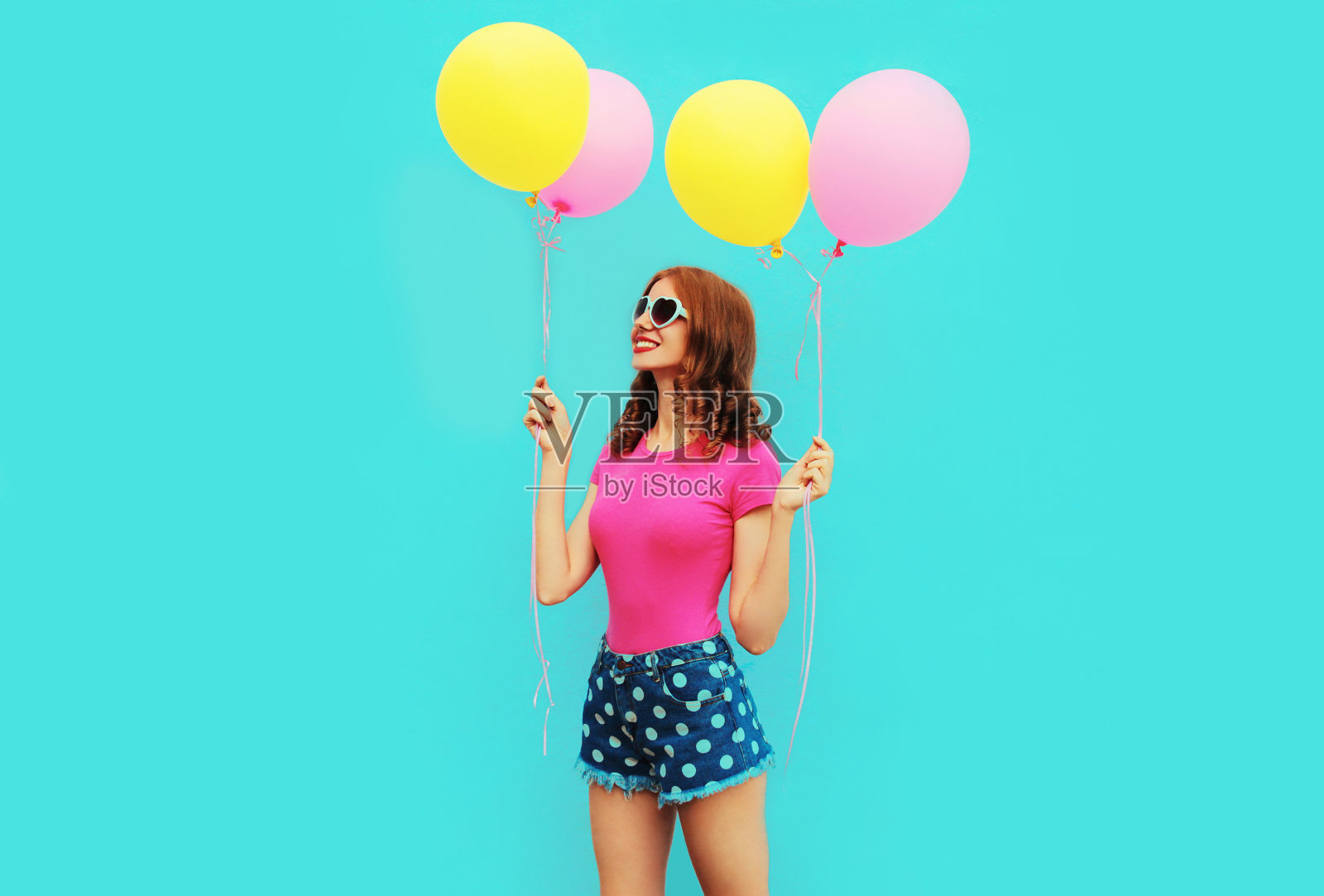 夏天彩色的愉快的图像，快乐的年轻女子与黄色粉红色的气球，穿着短裤和草帽在蓝色的墙壁背景照片摄影图片