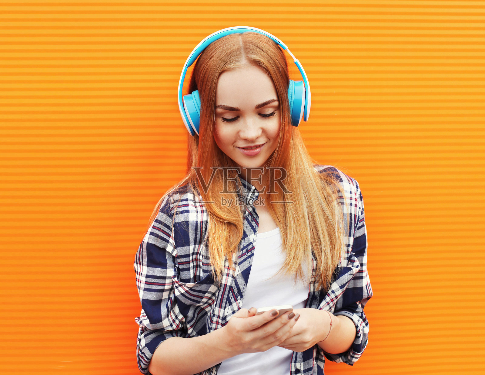 年轻女子的肖像与手机在无线耳机听音乐穿着休闲衣服在橙色的背景照片摄影图片