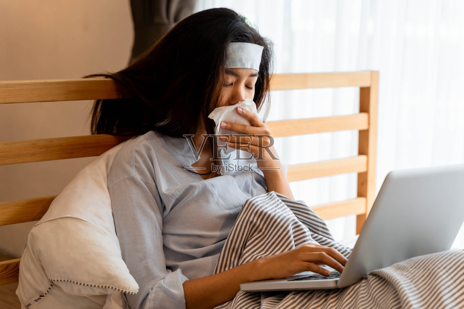20多岁的年轻亚洲女性在家里卧室用凝胶片降温的肖像。虽然她生病了，但还是在家工作。高烧、流感、流感、偏头痛、冠状病毒症状照片摄影图片
