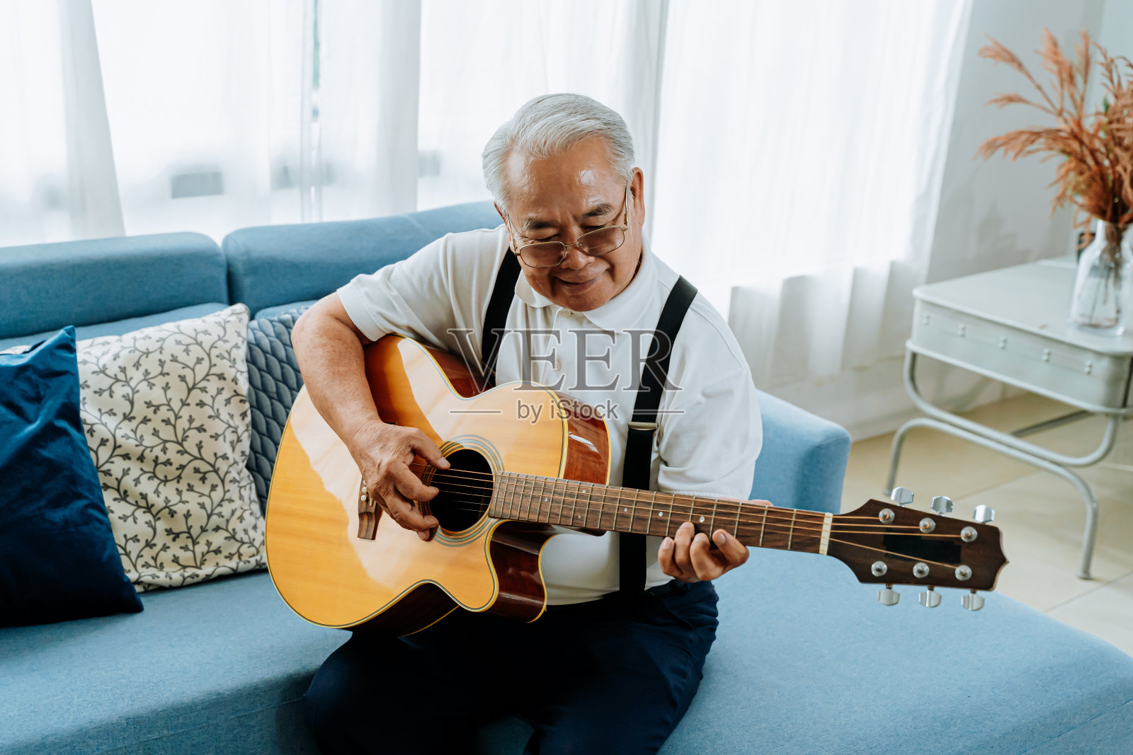 亚裔老人坐在沙发上弹奏原声吉他。快乐的微笑老人唱着吉他享受。在家享受退休生活。照片摄影图片