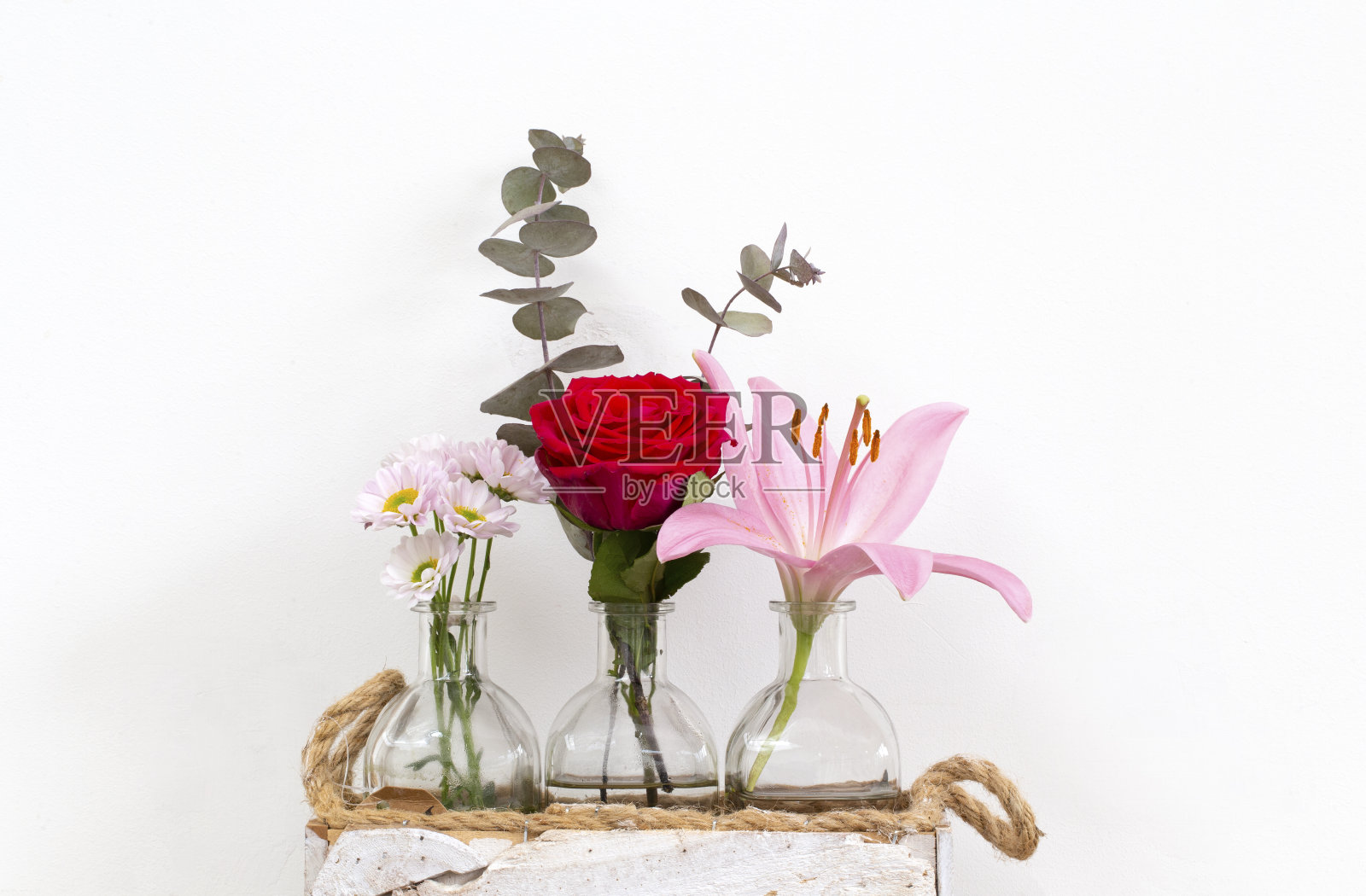 白色墙壁旁边的小玻璃花瓶里装着夏天的鲜花，鲜亮的红玫瑰、粉色的百合和雏菊。现代装饰家里照片摄影图片