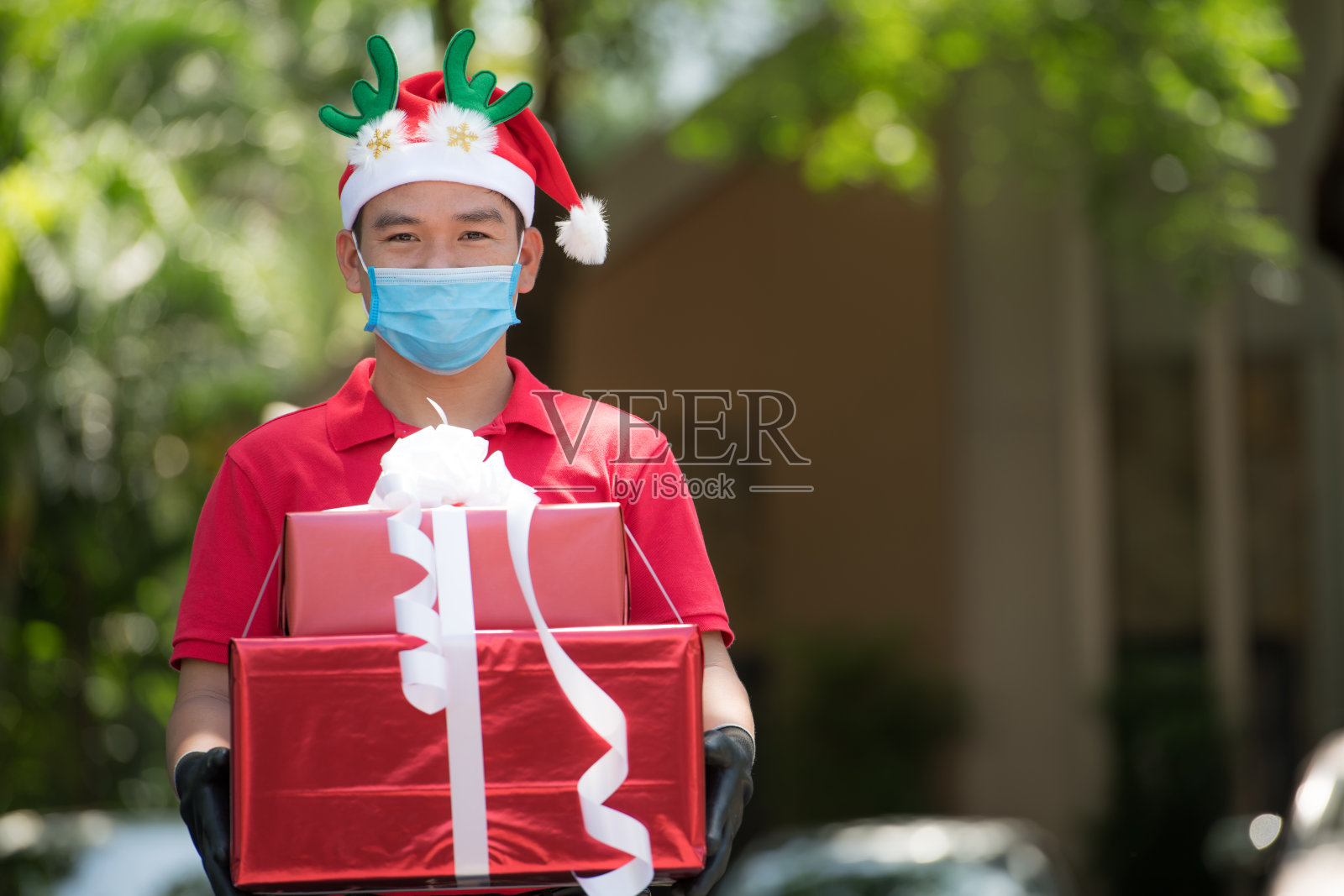 2019冠状病毒肺炎疫情期间，亚洲快递员身穿红色制服，戴着口罩和手套，戴着圣诞帽，在圣诞节期间派送礼物和礼品盒照片摄影图片