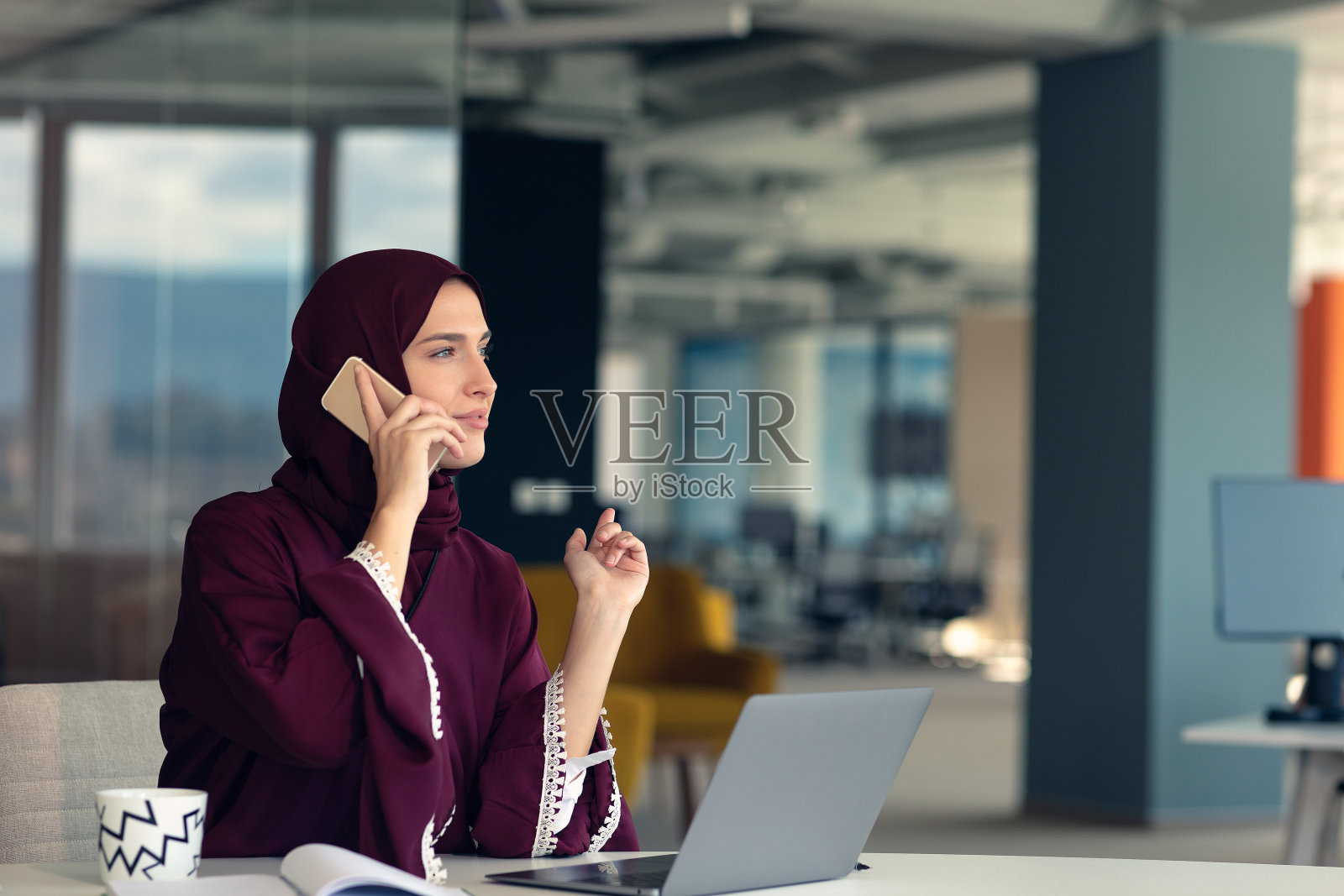 快乐的穆斯林女商人戴着头巾在办公室工作。微笑的阿拉伯妇女使用笔记本电脑和智能手机工作。照片摄影图片