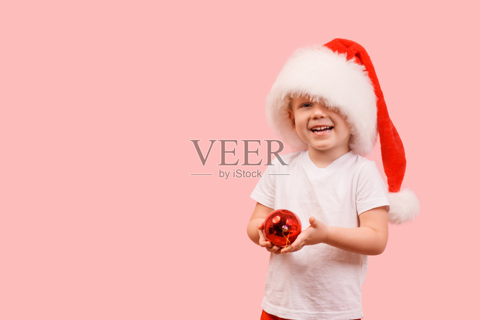 3岁的小孩戴着圣诞老人的帽子站着，举着新年球。工作室拍摄的概念，明信片的粉红色背景与地方的文字和祝贺。照片摄影图片