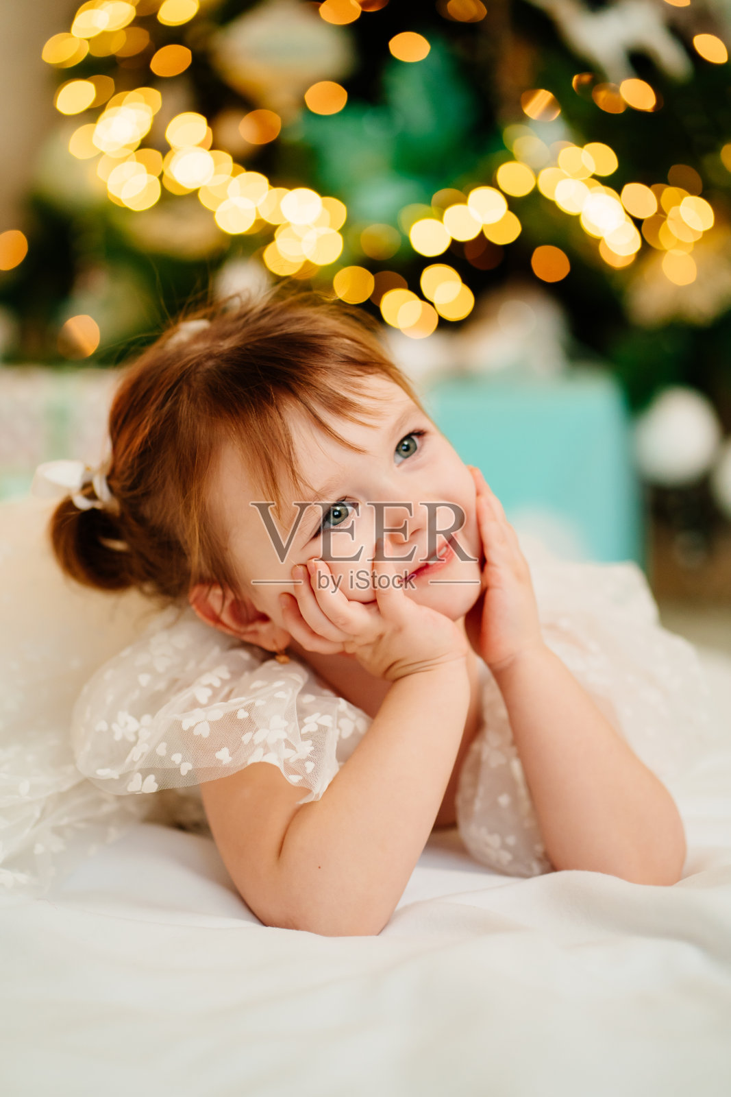 一个穿着白色连衣裙的小女孩躺在房子的圣诞树下。照片摄影图片