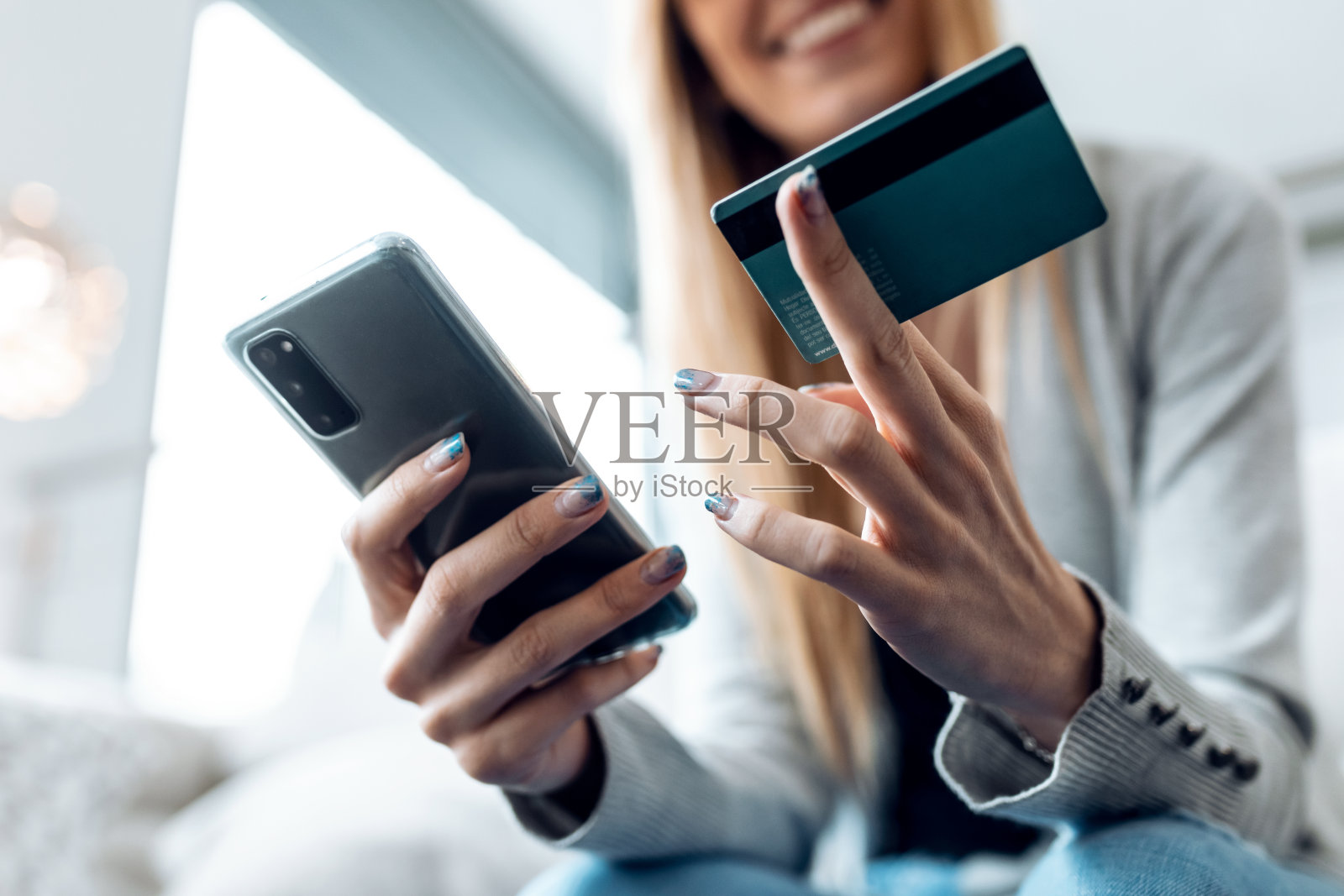 微笑的年轻女子坐在家里的沙发上用她的信用卡和智能手机在网上支付东西。照片摄影图片