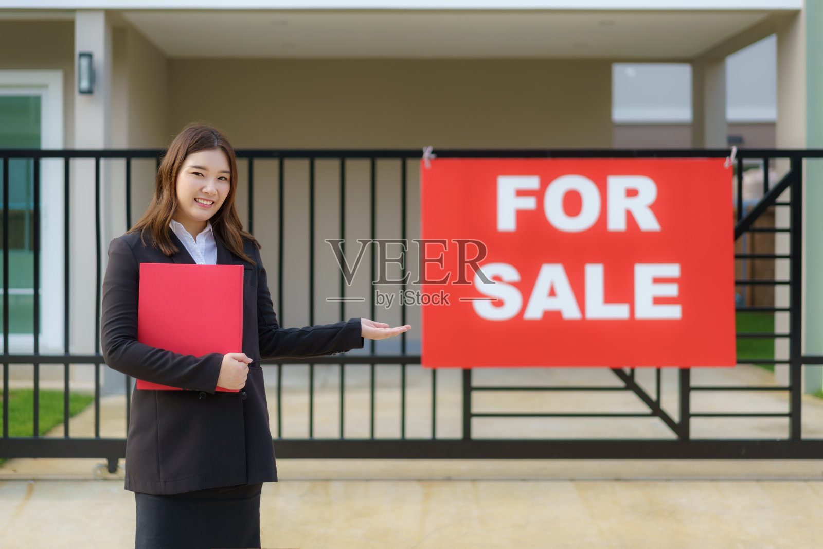 亚洲女性的房地产经纪人站在房前，门阶上挂着销售标志，宣布有兴趣的各方正在联系。照片摄影图片