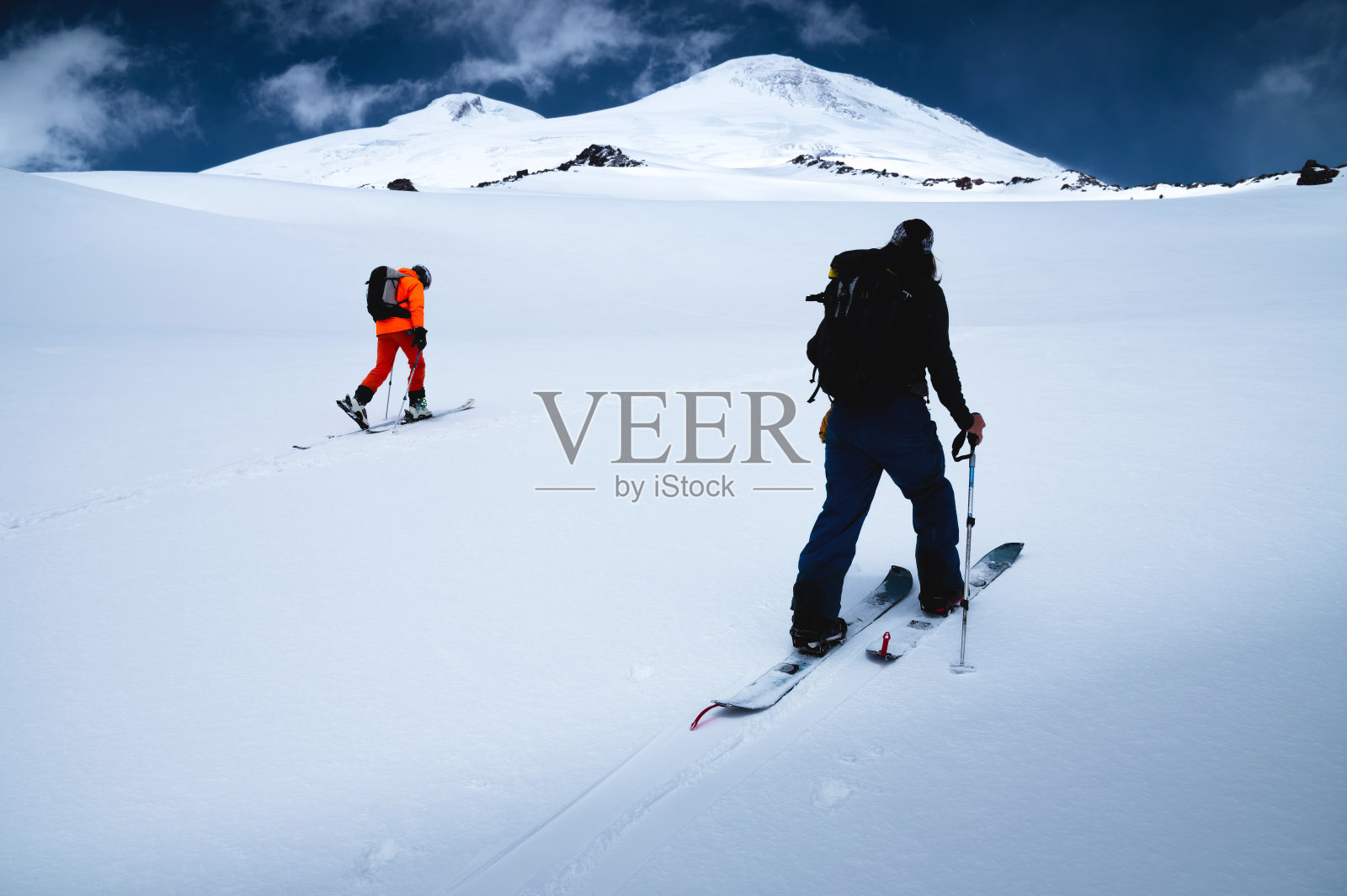 在一次滑雪之旅中，两个免费乘车的滑雪者爬上了刚下过的雪。厄尔布鲁斯山的顶峰和背景上的蓝天照片摄影图片