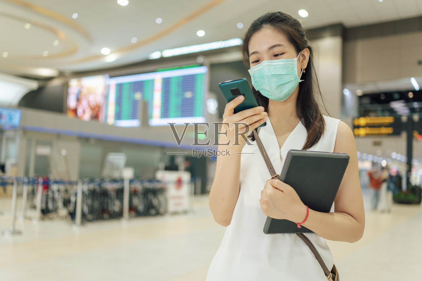 在机场候机区，年轻的亚洲成年商务女性坐在候机厅前，一边等待登机，一边在平板电脑上查看工作或邮件照片摄影图片