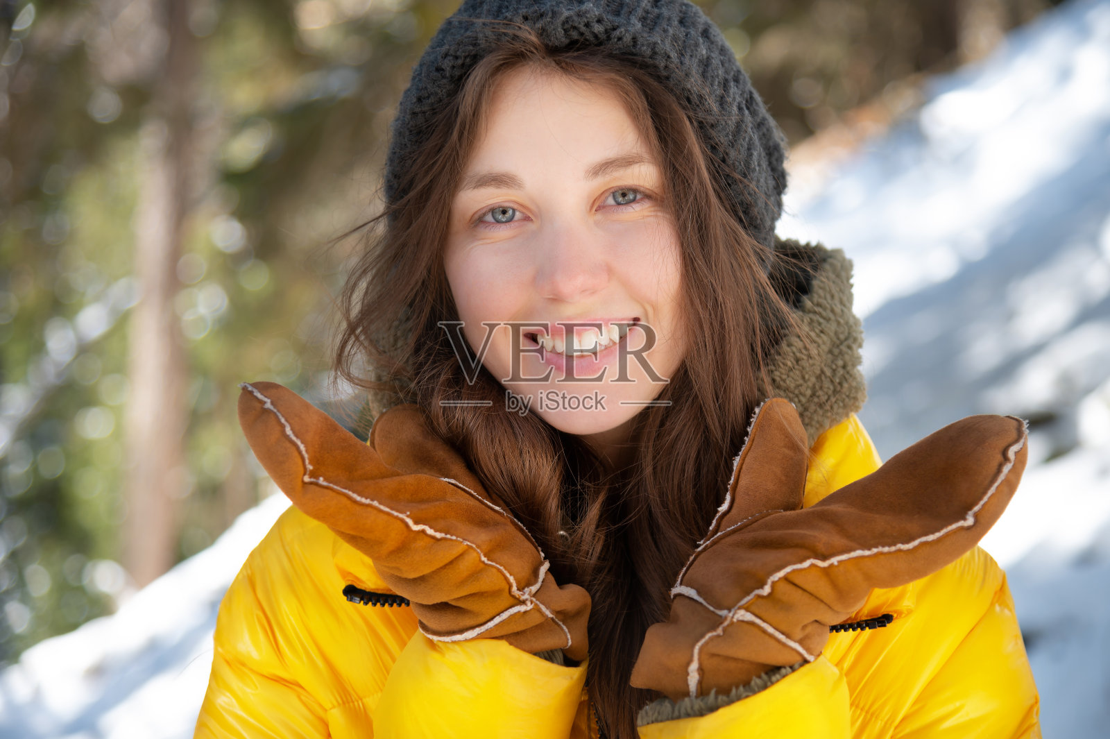 在一个阳光明媚的日子里，一名年轻漂亮的白人女子身穿冬季黄色羽绒服，头戴针织帽，微笑地看着镜头，背景是白雪皑皑的针叶林照片摄影图片