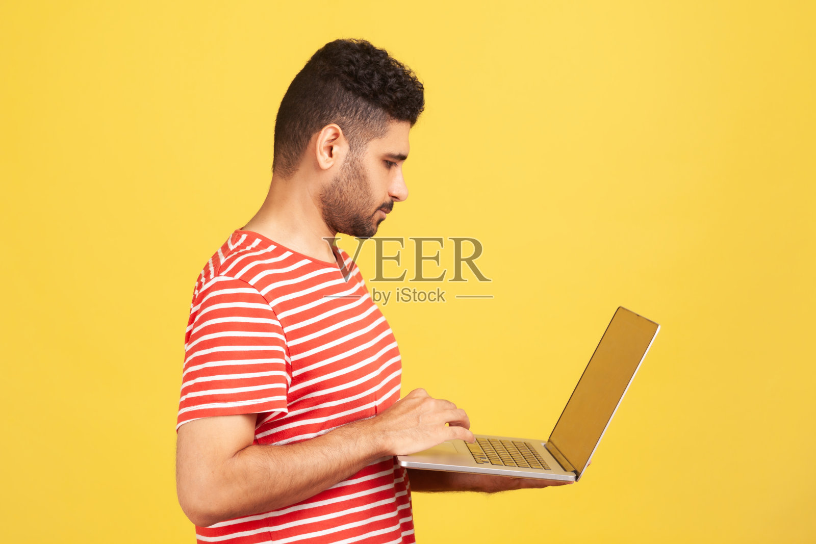 侧影肖像严肃专注的男子胡子在红条纹t恤工作在笔记本电脑上，站在键盘上打字，远程工作照片摄影图片