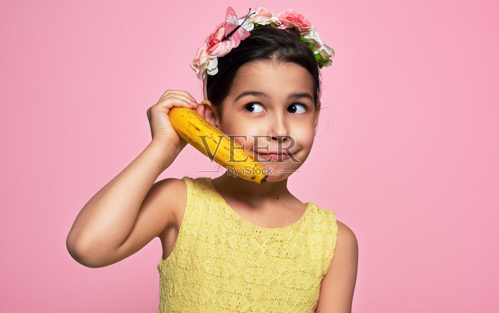 一个有趣的小女孩拿着香蕉，像手机一样，孤立在粉红色的背景下。一个穿着黄色连衣裙的快乐的孩子假装对着香蕉说话，就像对着手机一样。照片摄影图片
