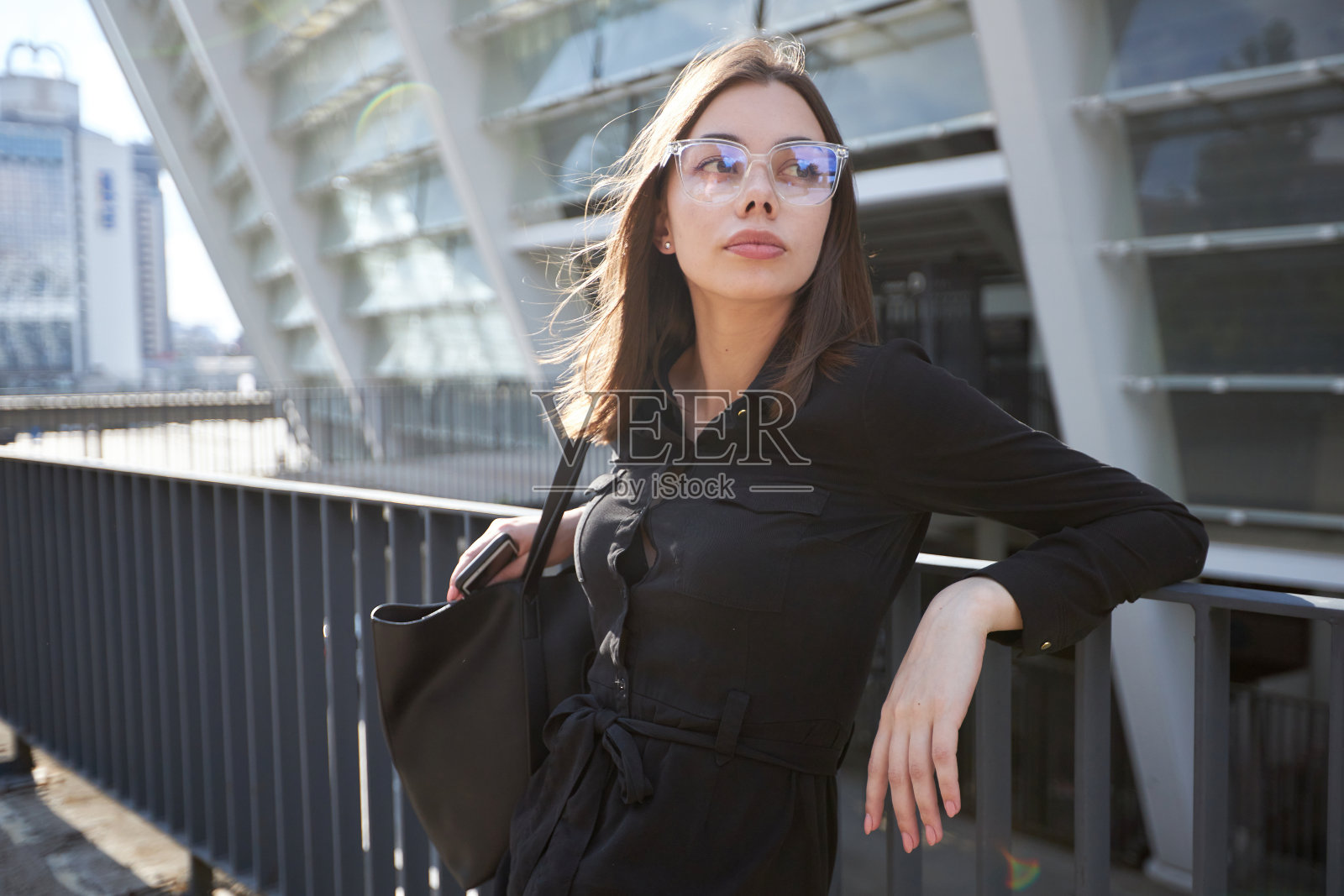 商业妇女肖像在城市街道在日落。性感的黑发，穿着黑色的裙子，戴着眼镜和黑色的皮包照片摄影图片