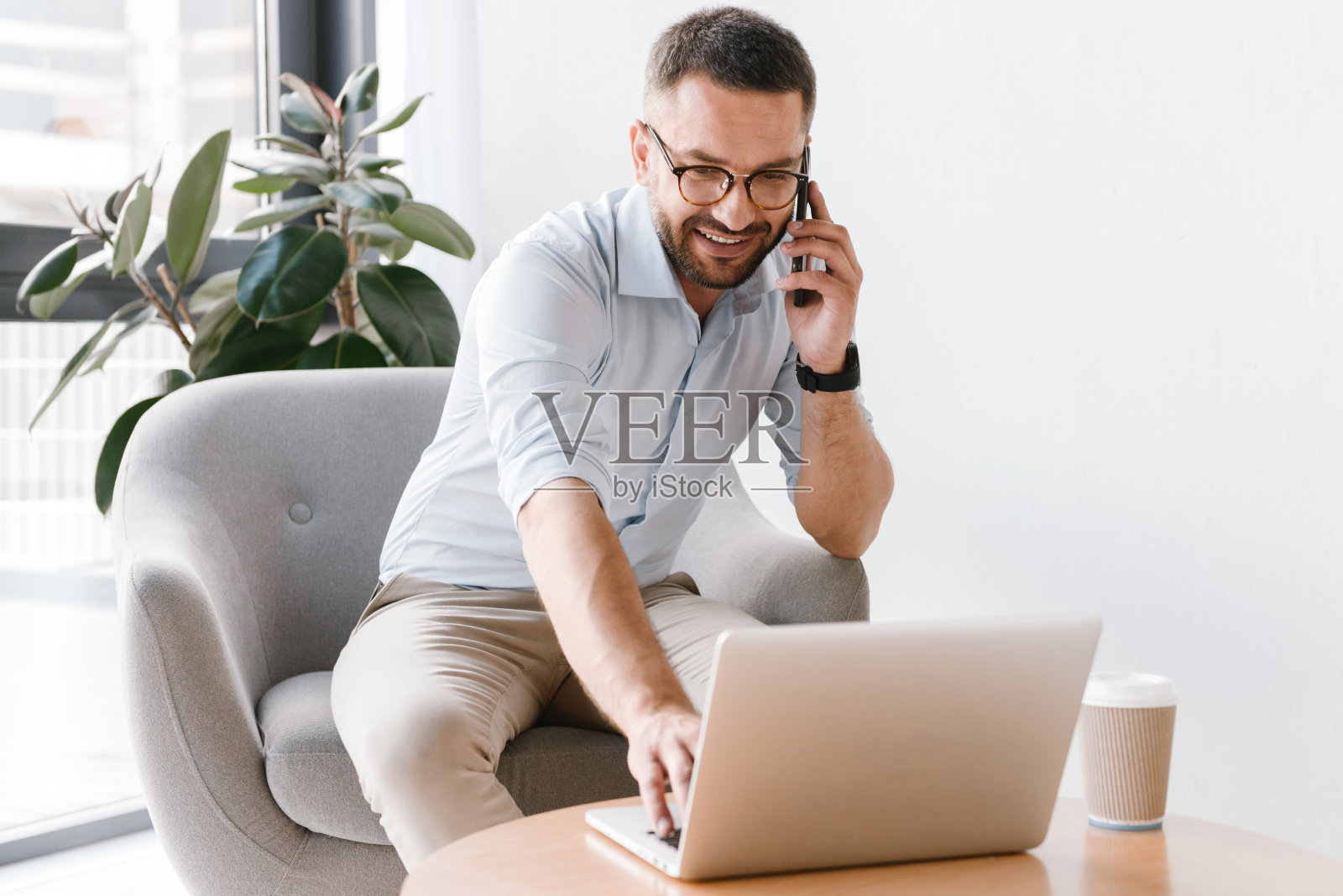 照片中的欧洲男人30多岁，穿着白衬衫坐在扶手椅上，使用笔记本电脑，同时有业务电话照片摄影图片