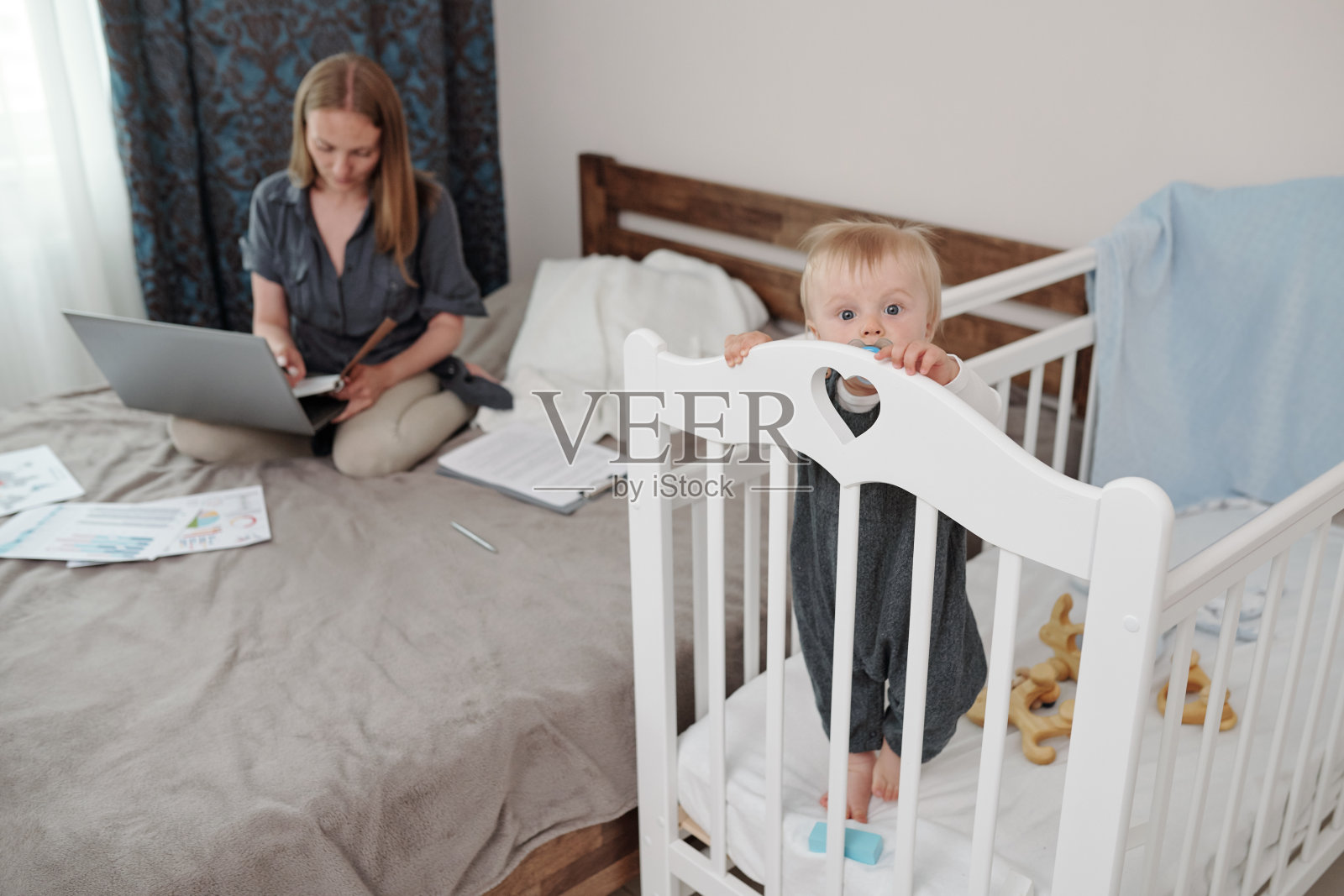 母亲工作时，婴儿站在婴儿床上照片摄影图片