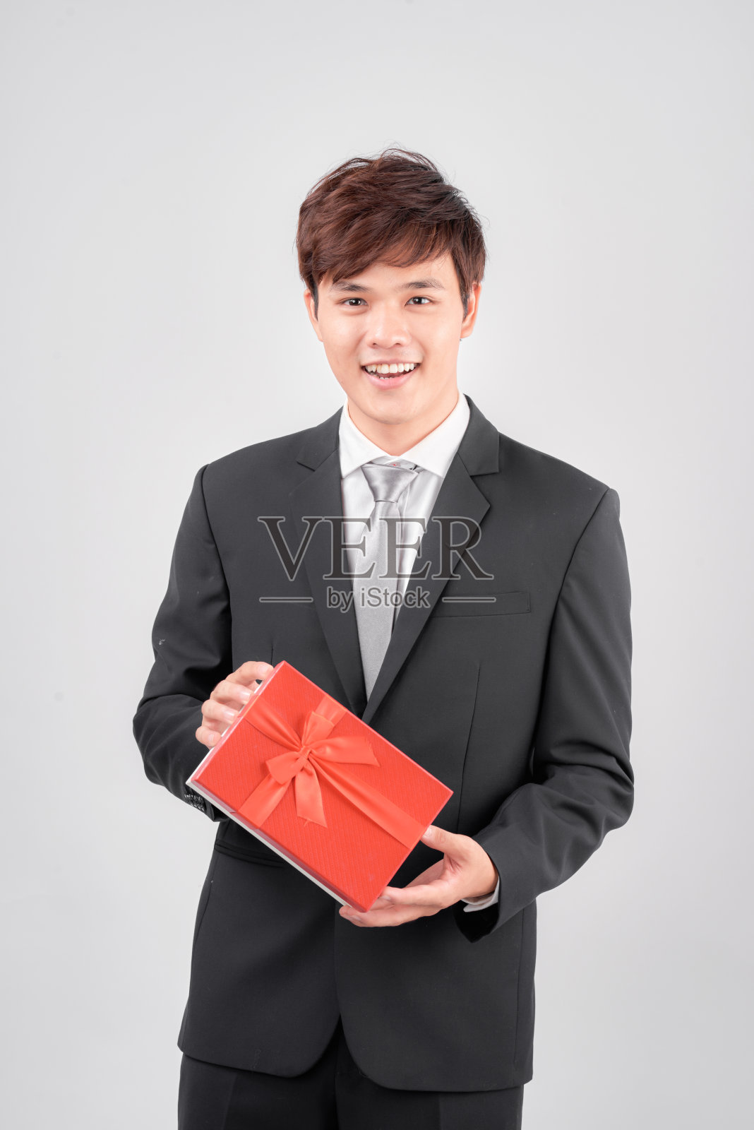 身着西装的帅气青年男子手拿红色礼品盒，站在白色背景下，望着镜头微笑。照片摄影图片