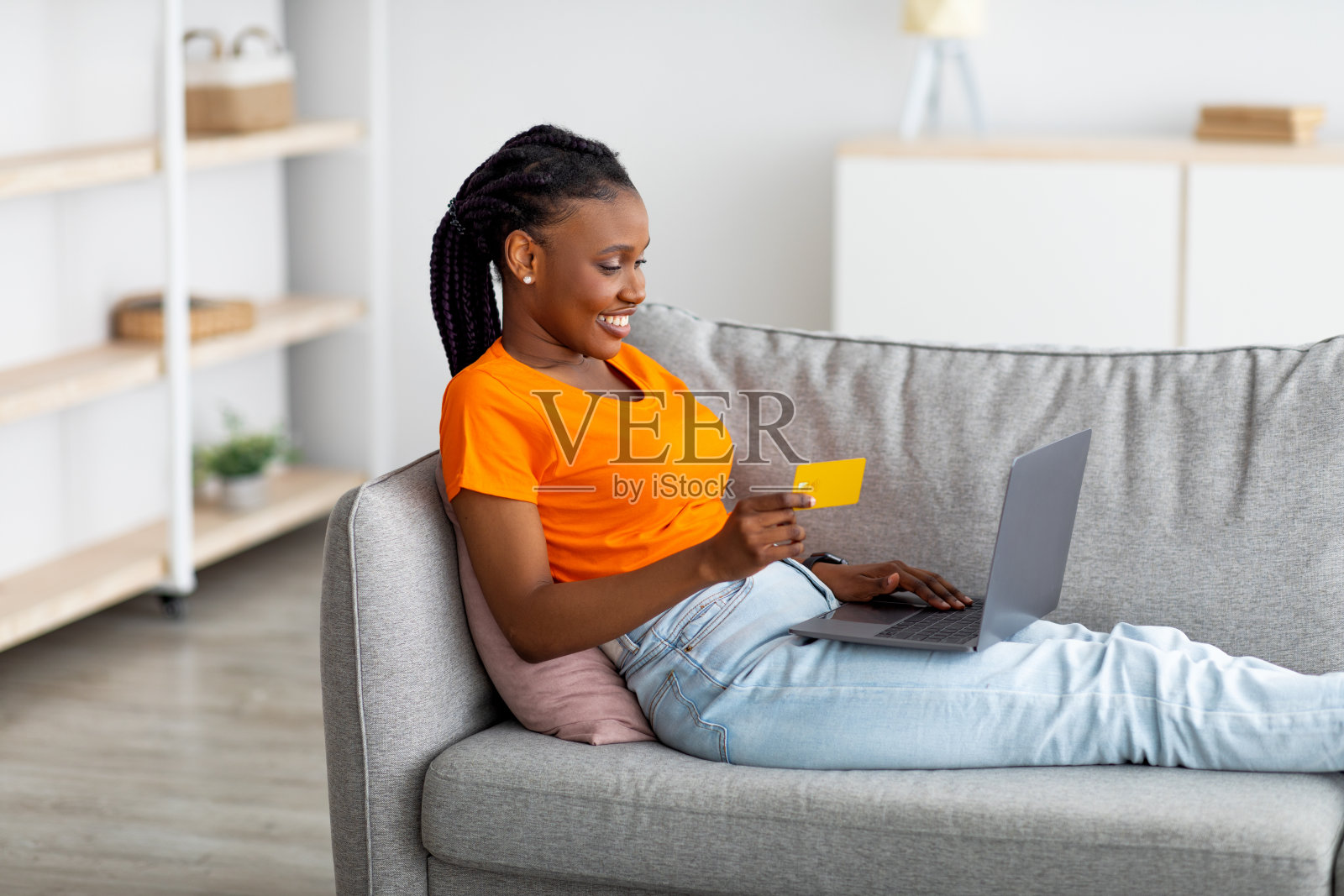 年轻的黑人女性坐在沙发上，使用笔记本电脑和信用卡，在家里使用网上购物或银行网站照片摄影图片