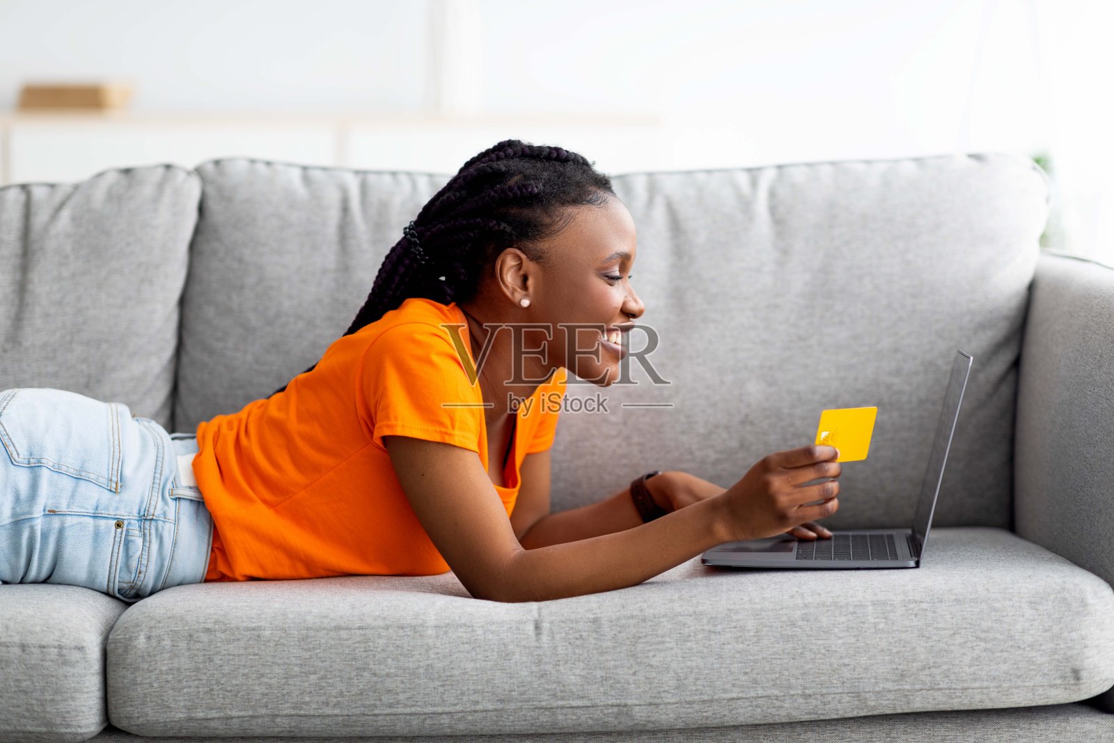 千禧一代的非洲女人在家网购，用笔记本电脑和信用卡，躺在沙发上，侧视照片摄影图片