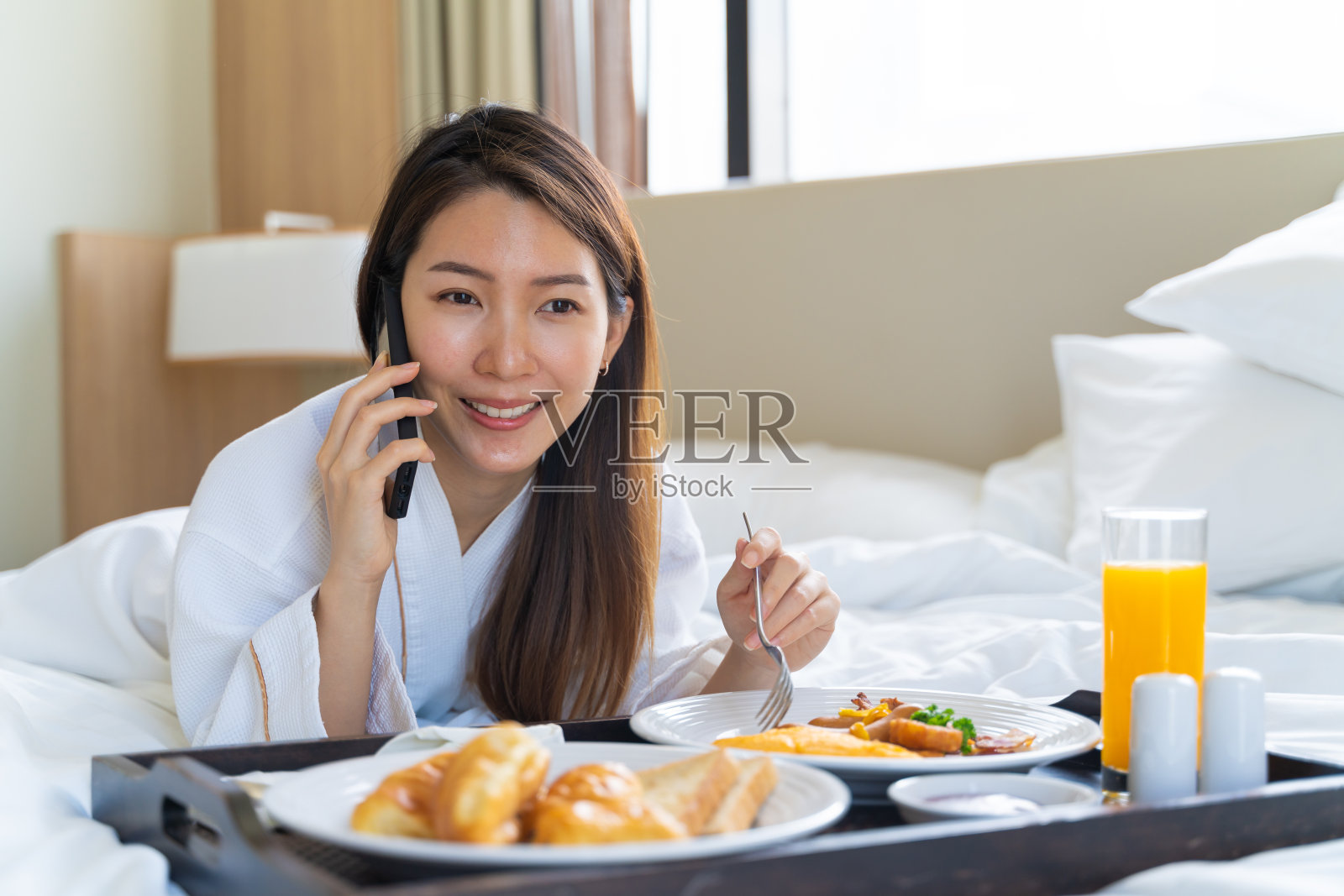 亚洲女人穿着浴袍在卧室的床上一边吃早餐一边讲电话。照片摄影图片