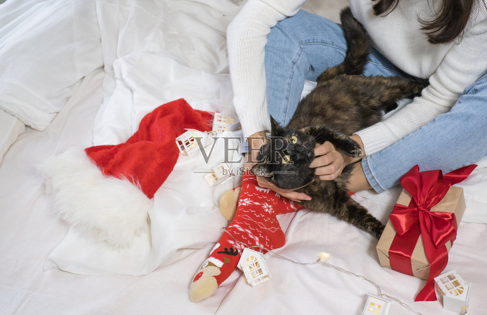 一个穿着明亮的新年袜子的女孩正抚摸着白床上的黑猫，手里拿着圣诞礼物和圣诞老人的帽子。照片摄影图片